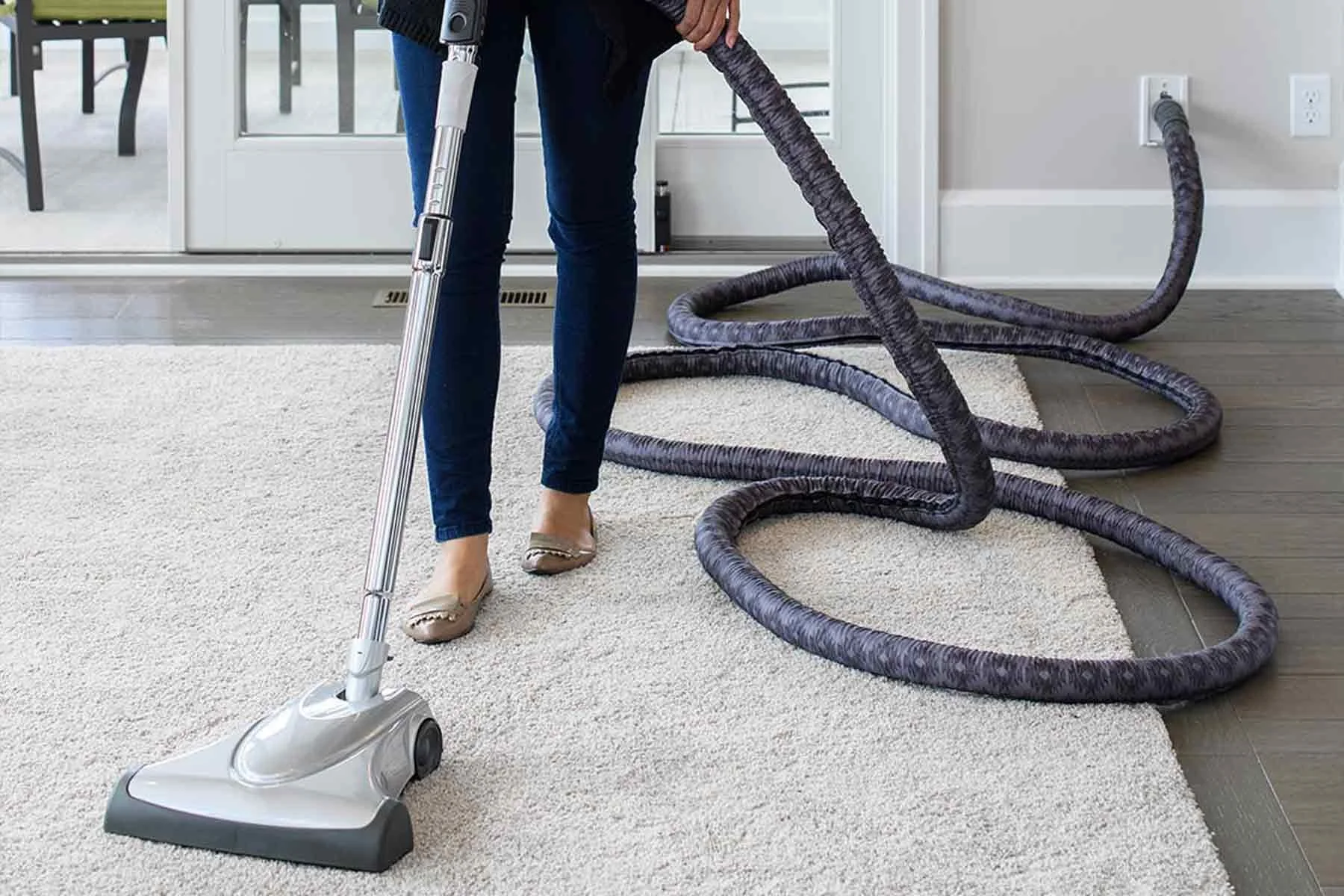 Person vacuuming a room with a central vacuum system, with a flexible hose and a vacuum cleaner attachment, on a beige carpet next to a white rug, in a room with a glass door and electrical outlets.