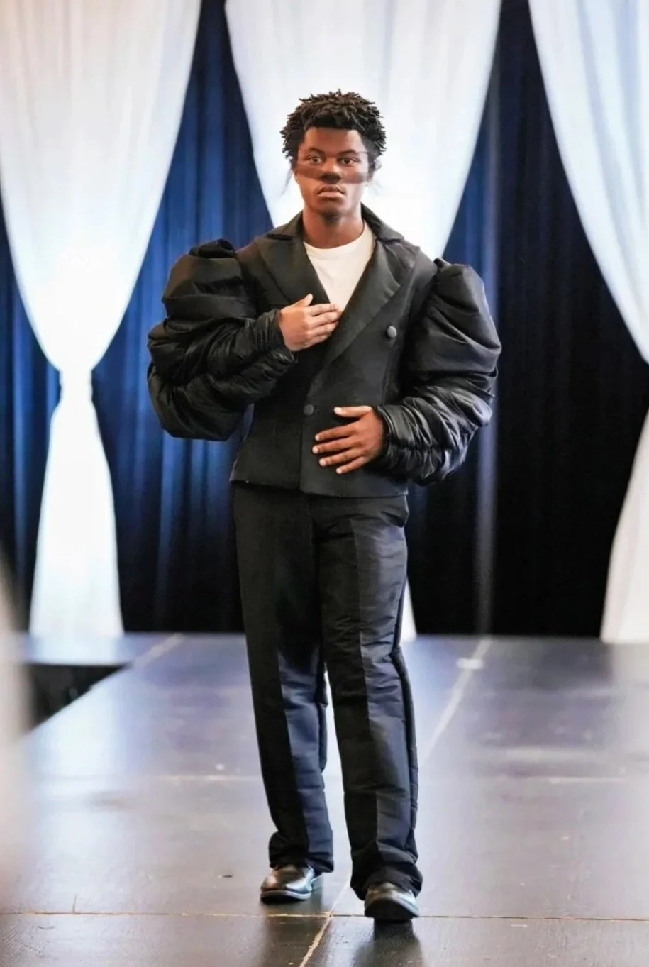 A young man standing on a stage with black and white drapes in the background, wearing a black blazer with puffy sleeves, black pants with a stripe down the side, and holding his right hand over his heart, appearing to be in a formal or ceremonial setting.