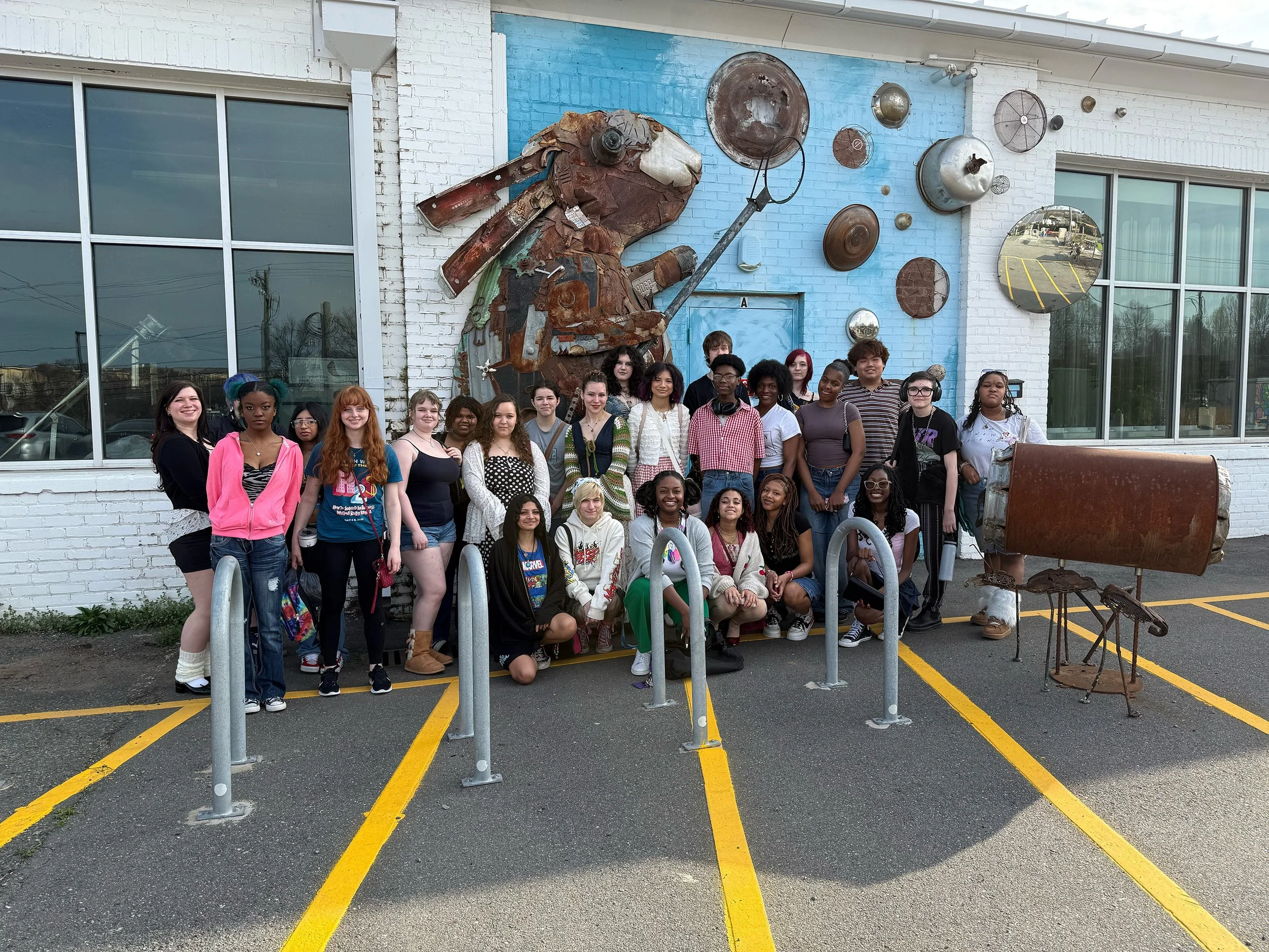 Group of diverse young people standing in front of a building with a large mural of a dog made of metal scraps, and various metal objects mounted on the wall. The ground is marked with yellow lines, and there are reflective windows on the building.