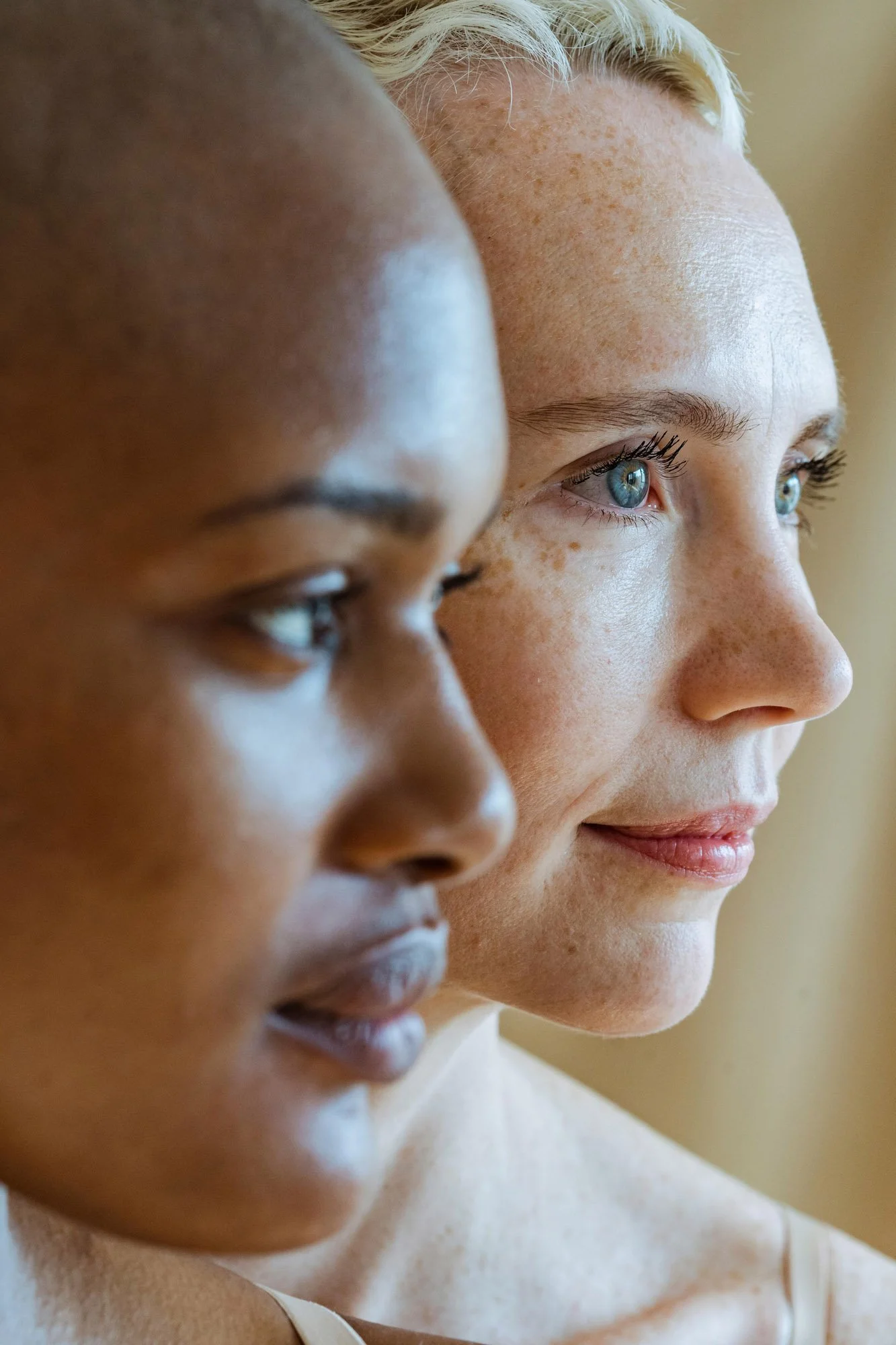 Deux femmes, l'une de peau foncée et l'autre de peau claire avec des tâches de rousseur, regardent dans la même direction, capturant un moment de concentration ou de réflexion.