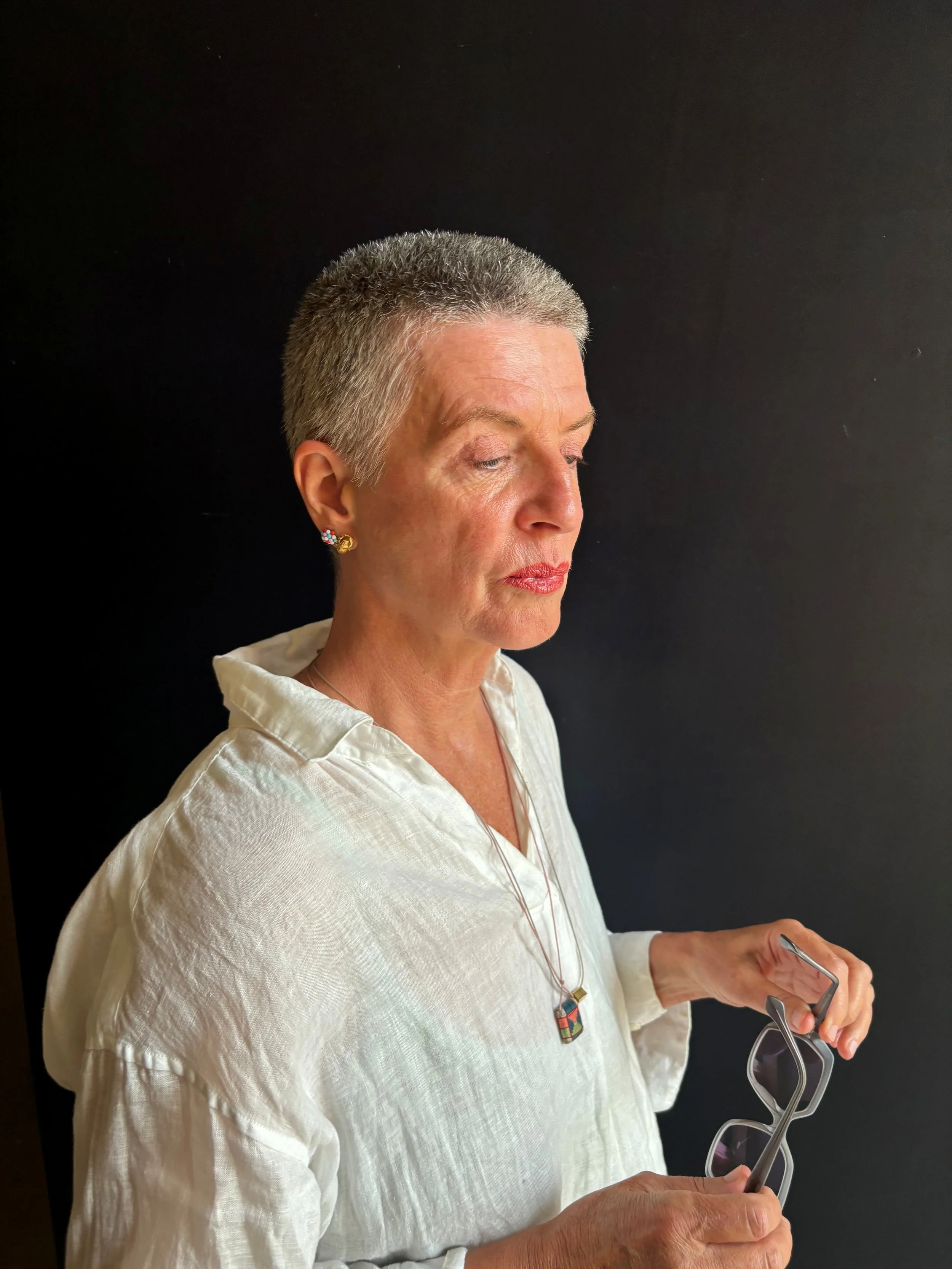An older woman with short gray hair, wearing a white blouse, earrings, and a colorful necklace, standing against a black background. She is holding eyeglasses.