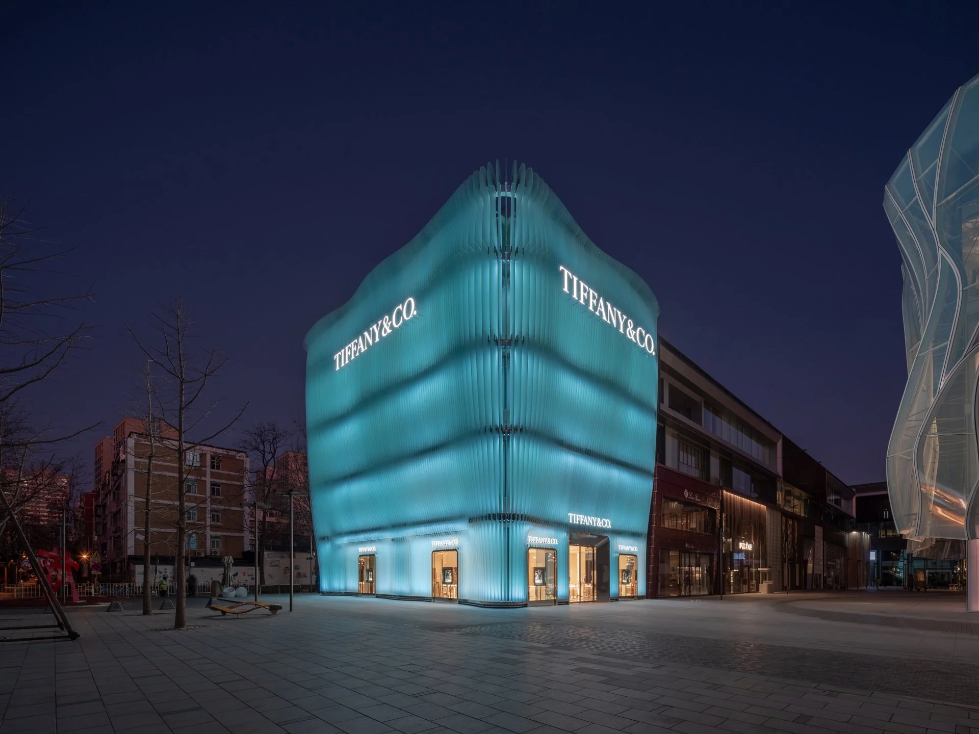 A modern, illuminated Tiffany & Co. store at night with a glowing, wavy facade in shades of blue and the brand's logo on the building.