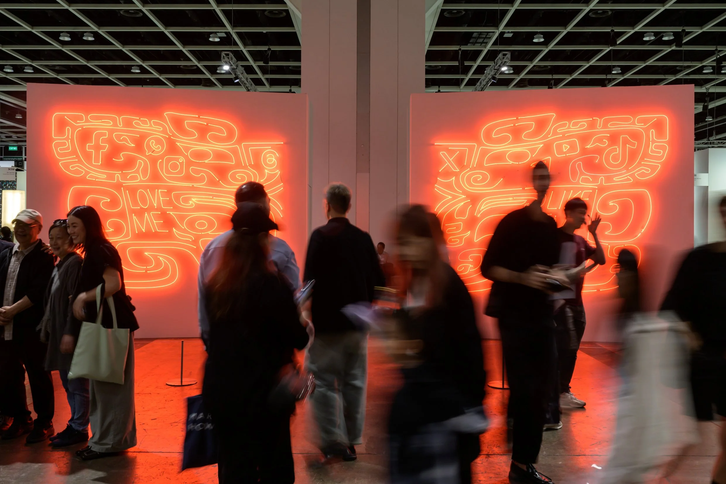 People walking in front of two large neon light art displays featuring social media icons, doodle-like designs, and words like 'LOVE', 'ME', and 'LIKE' at an indoor event.