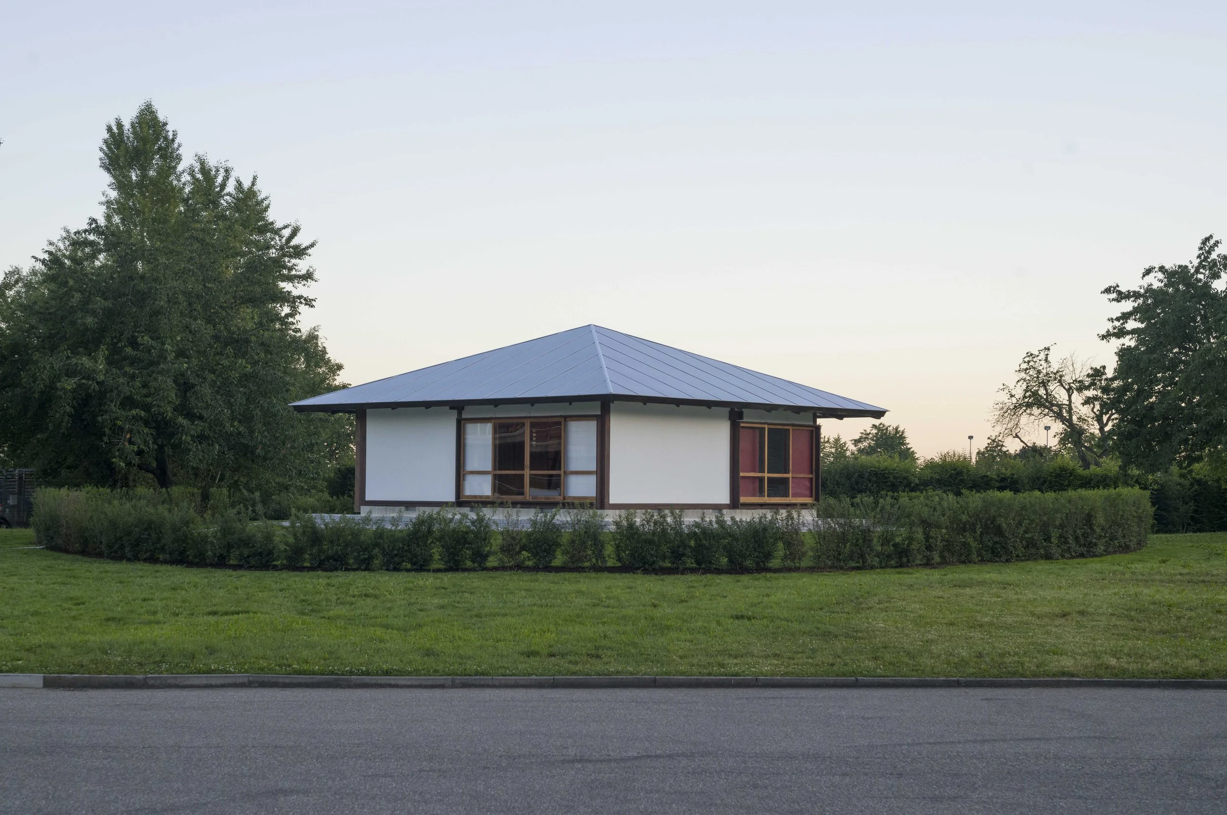 A small modern house with a metal roof and large windows, surrounded by green trees and bushes, with a grassy lawn and a paved road in the foreground, under a clear sky.