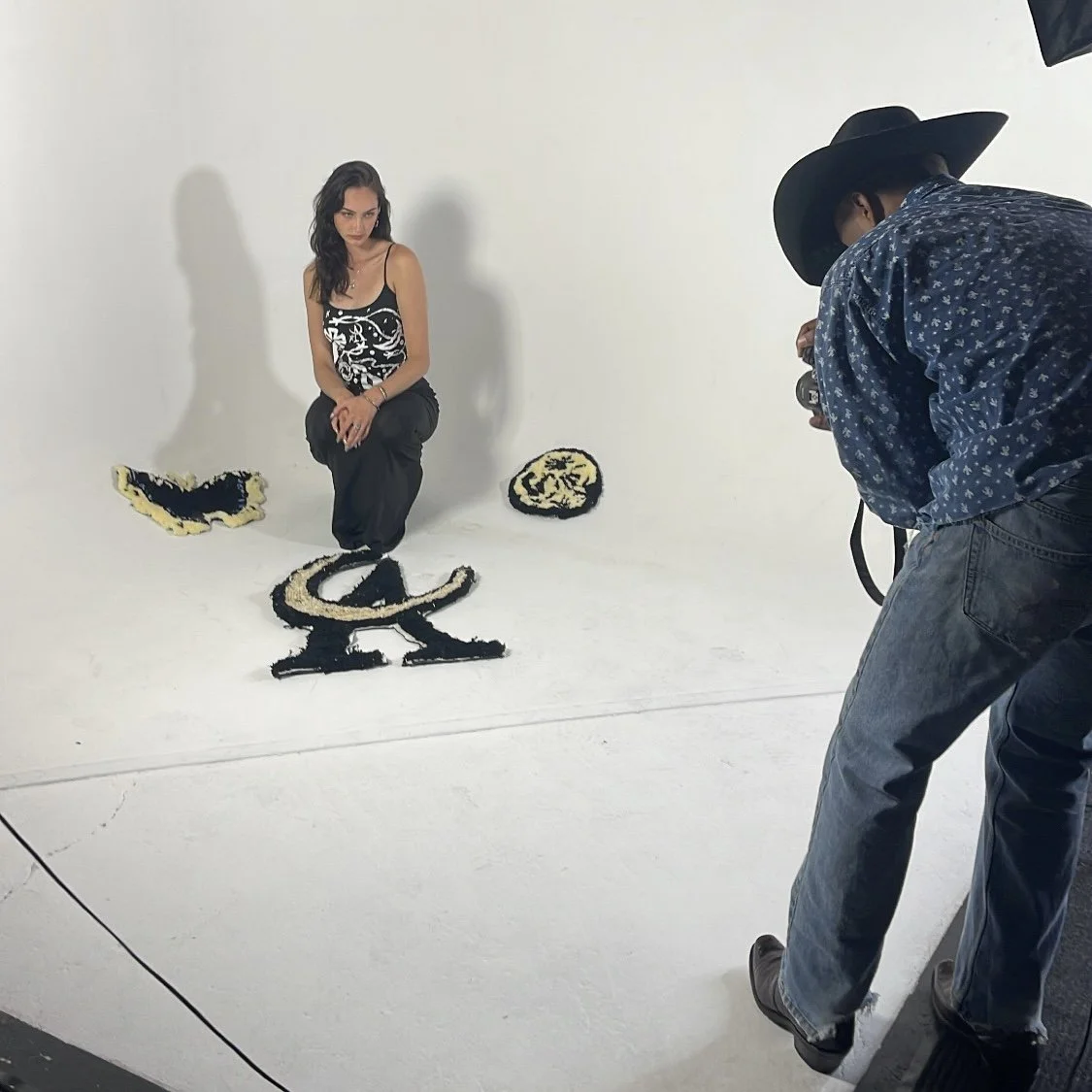 Photographer takes a picture of a woman sitting on her heels in a studio with a plain white background. The floor has artistic black and yellow shapes or symbols.