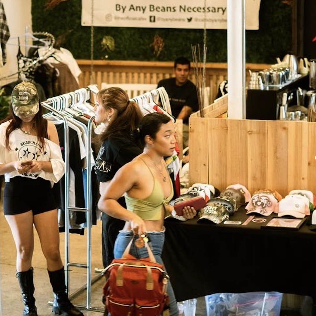 People browsing caps at an outdoor market stall with clothing racks and a wooden display, in front of a sign that says 'By Any Beans Necessary.'