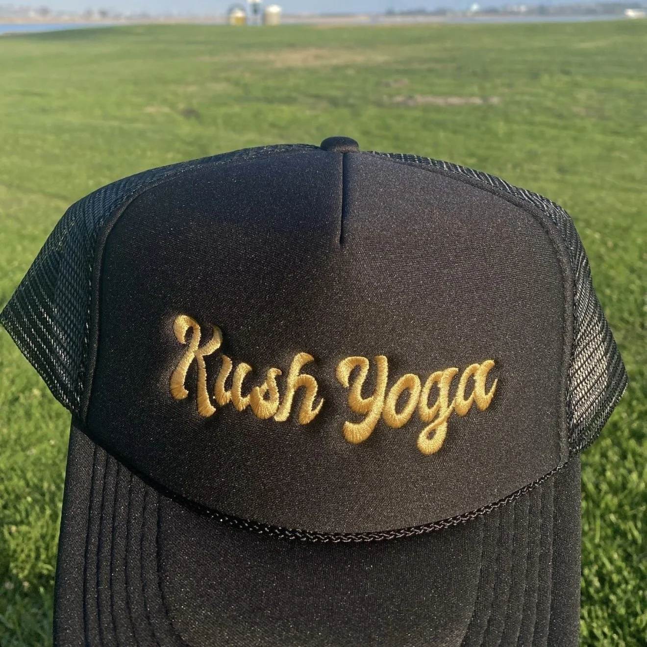 Black trucker hat with gold embroidery reading "Rush Yoga", placed on green grass with a blue sky and distant structures in the background.