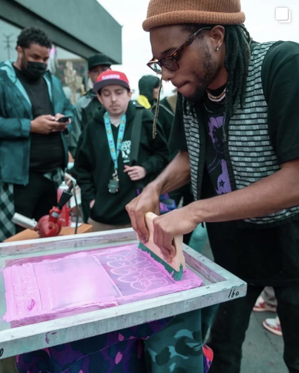 A man with sunglasses and a beanie in a brown color is screen printing a pink design onto fabric at an outdoor event, with several people observing.