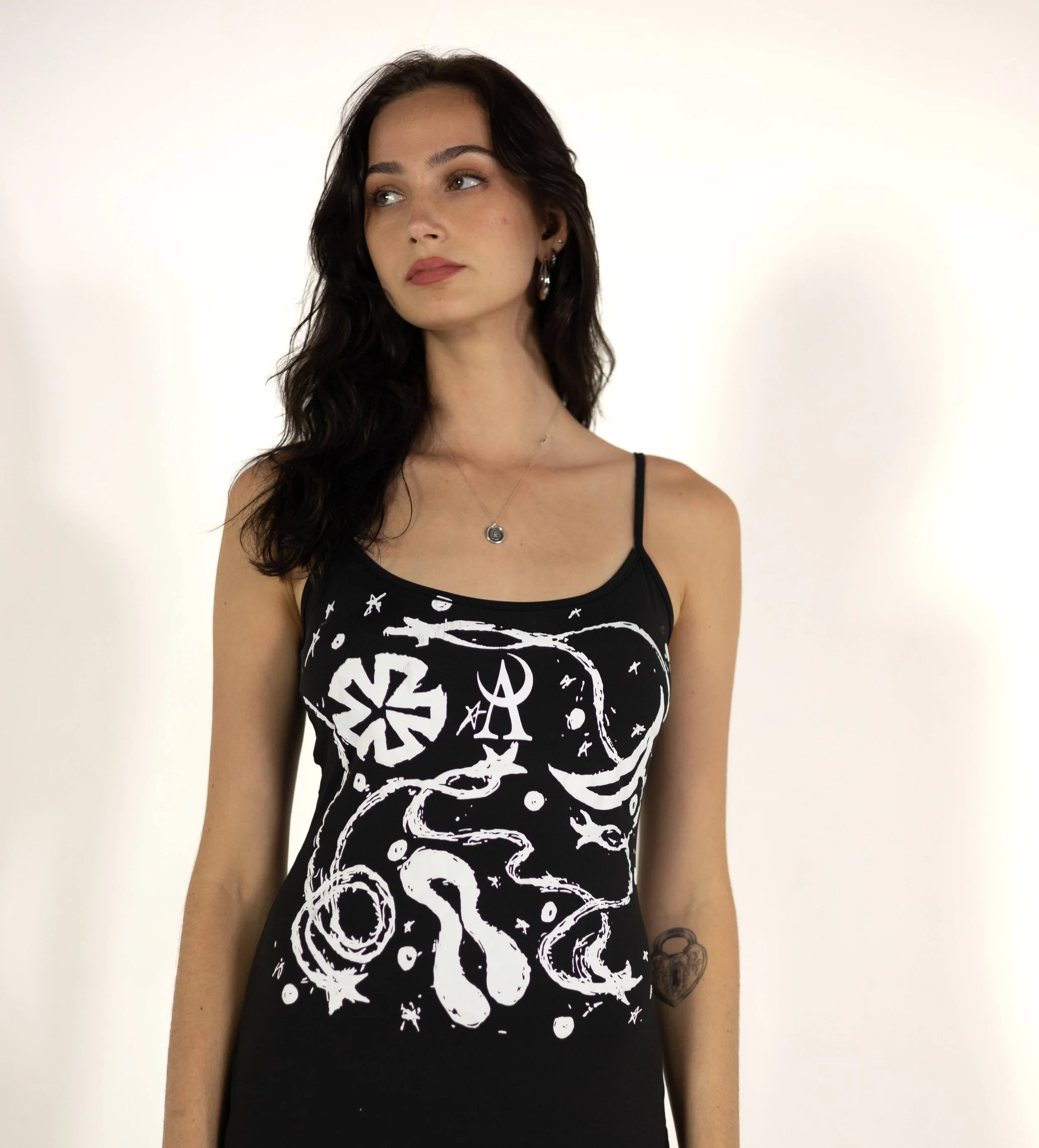 A woman with dark wavy hair and light skin, wearing hoop earrings and a necklace, stands against a plain white background. She is wearing a sleeveless black top with white graphic designs, including peace signs, stars, and various symbols.