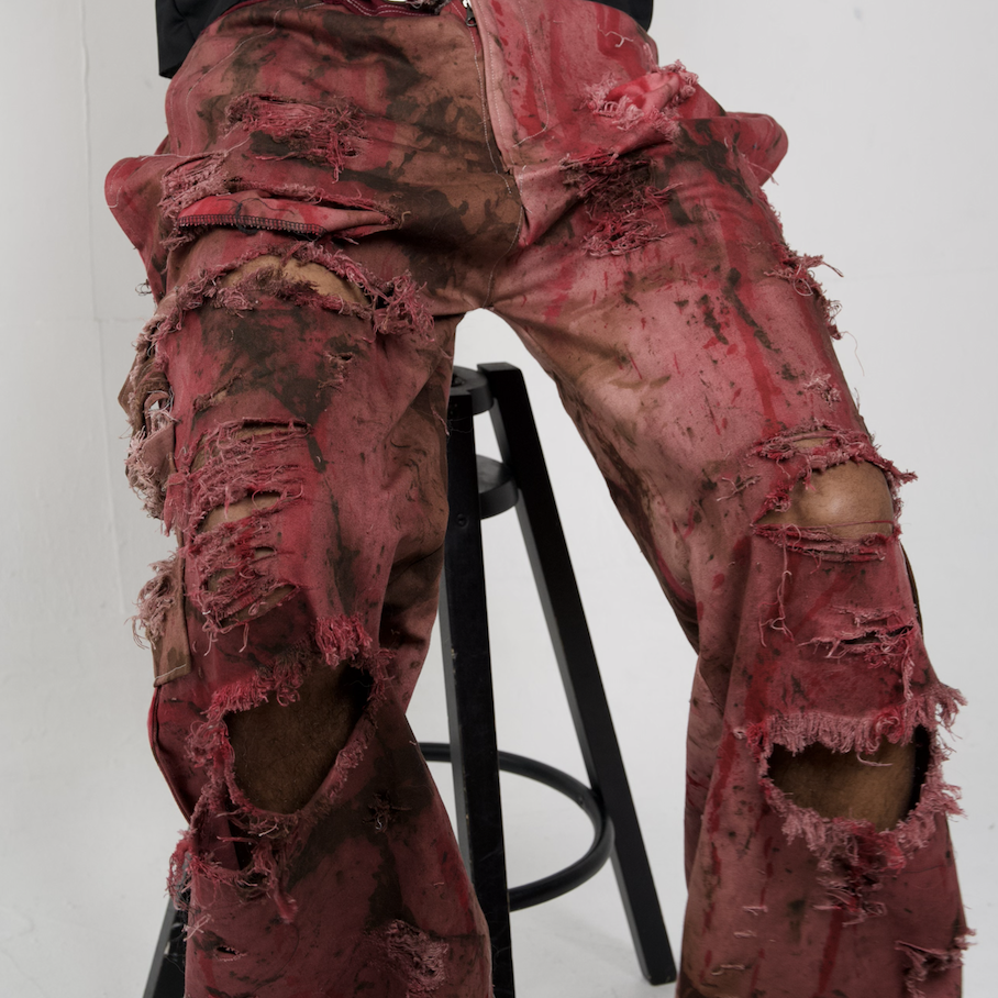 Person wearing heavily distressed, ripped, red and brown camouflage-patterned pants sitting on a black stool against a plain white background.