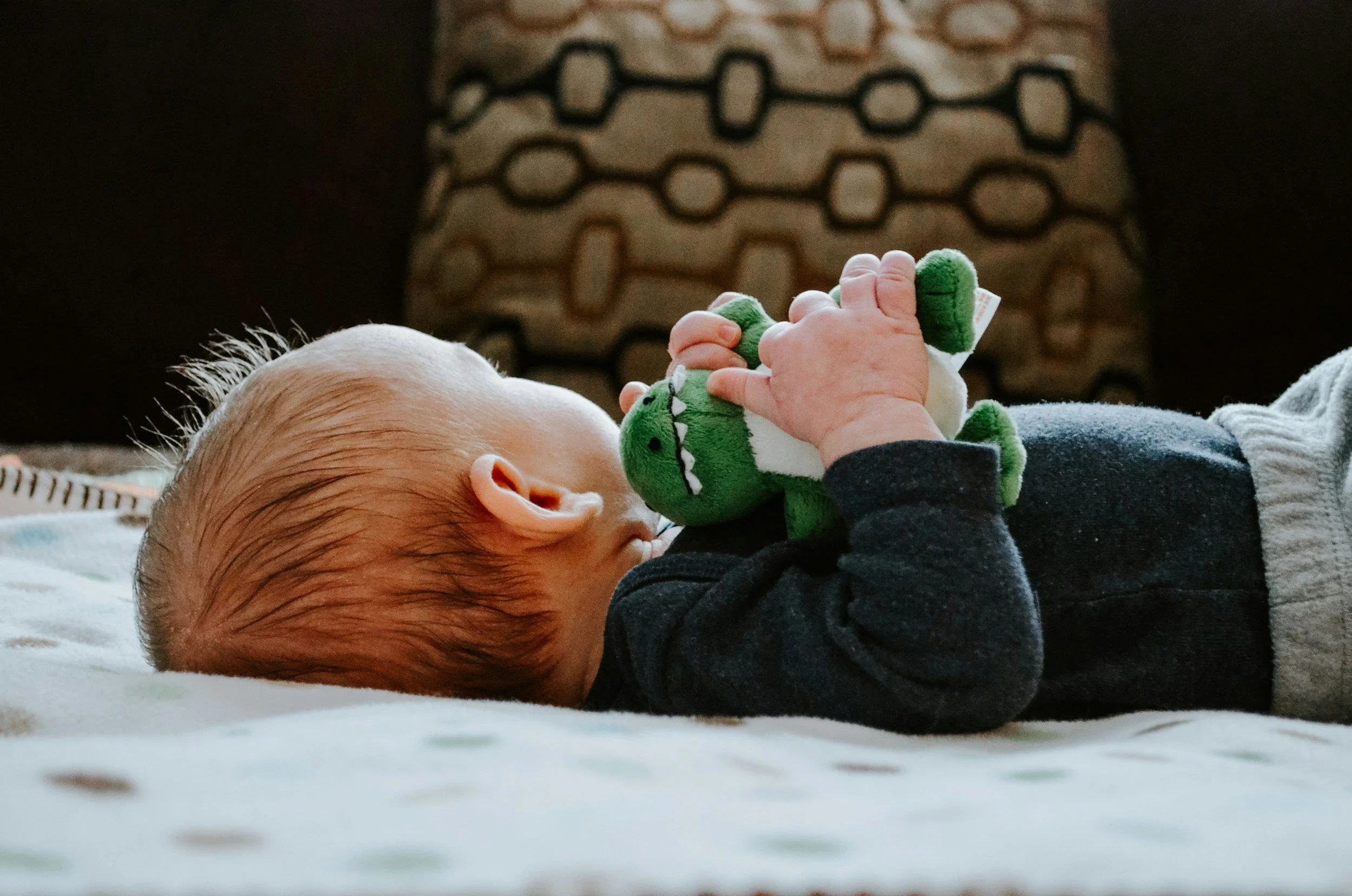 A sleeping baby with red hair lying on her stomach with a young blonde girl resting her head on her arm, both on a white bed.