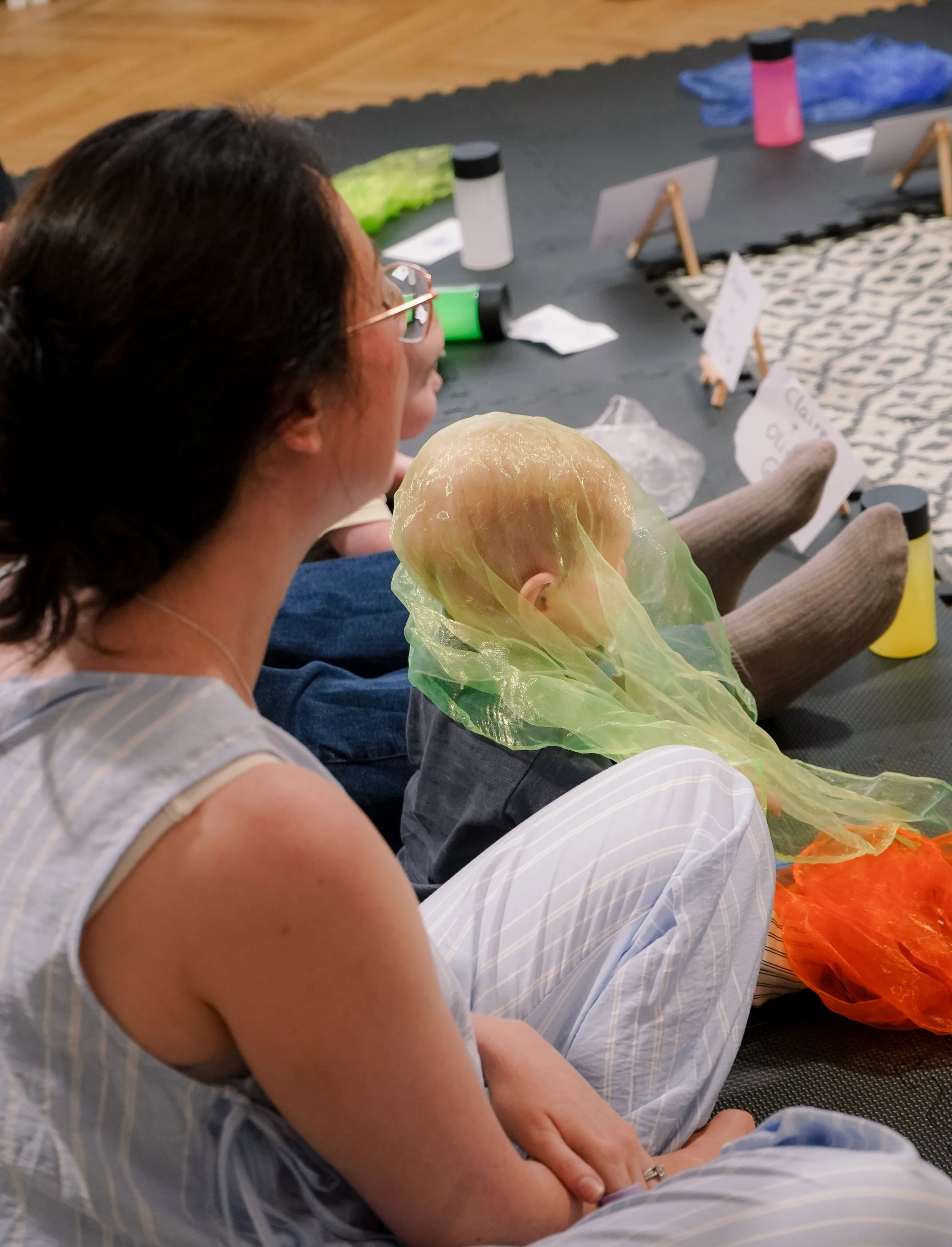 A woman with dark hair and glasses sitting on the floor, seen from the side, holding a baby wrapped in a yellow-green cloth. There are water bottles and signs on small easels on the floor behind them, with a patterned rug and wooden floor in the background.