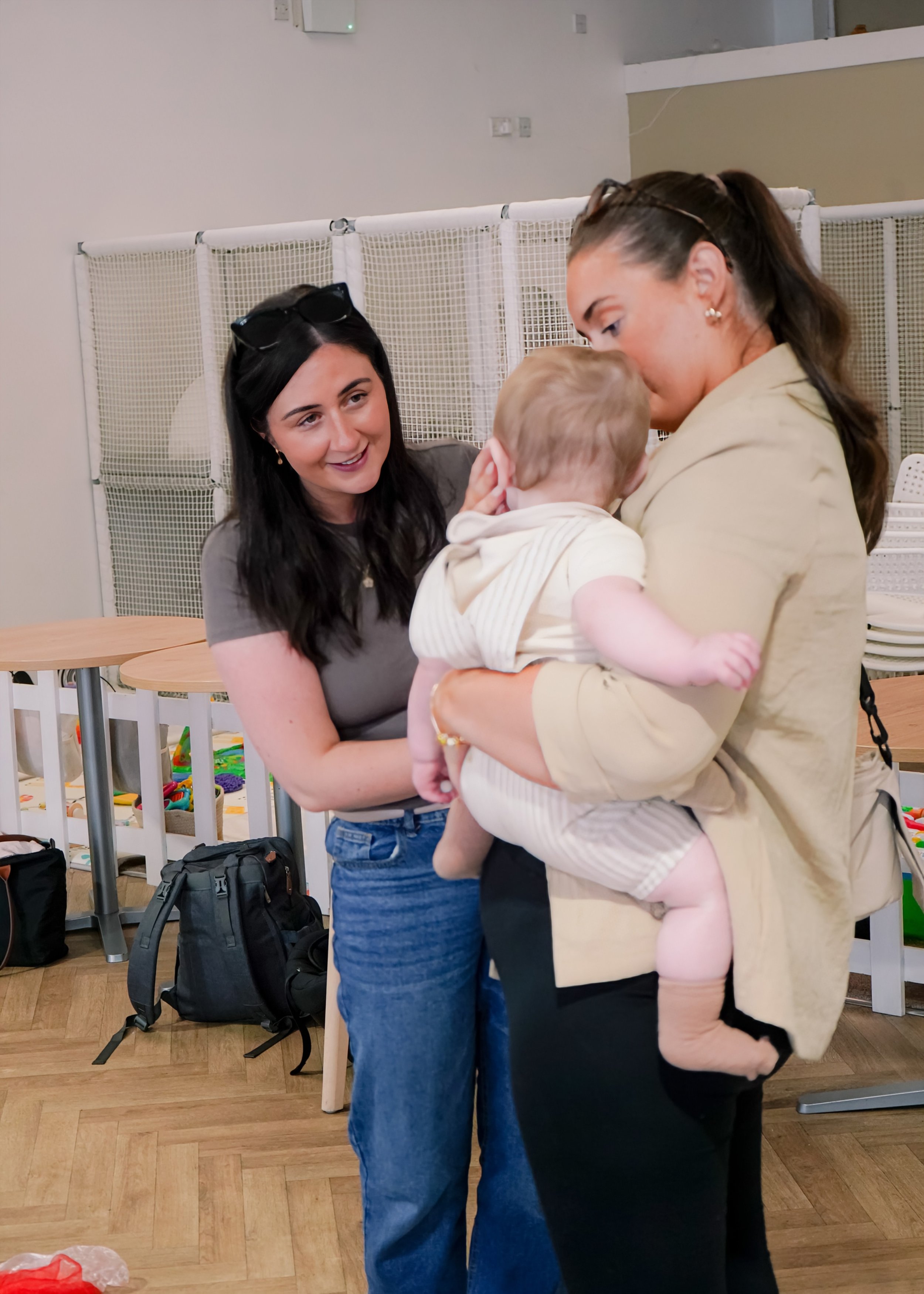 Two women and a young child inside a room. One woman is sitting, smiling at the woman holding the child. The woman holding the child is looking down at the child, who is touching her face. There are tables, chairs, and backpacks in the background.