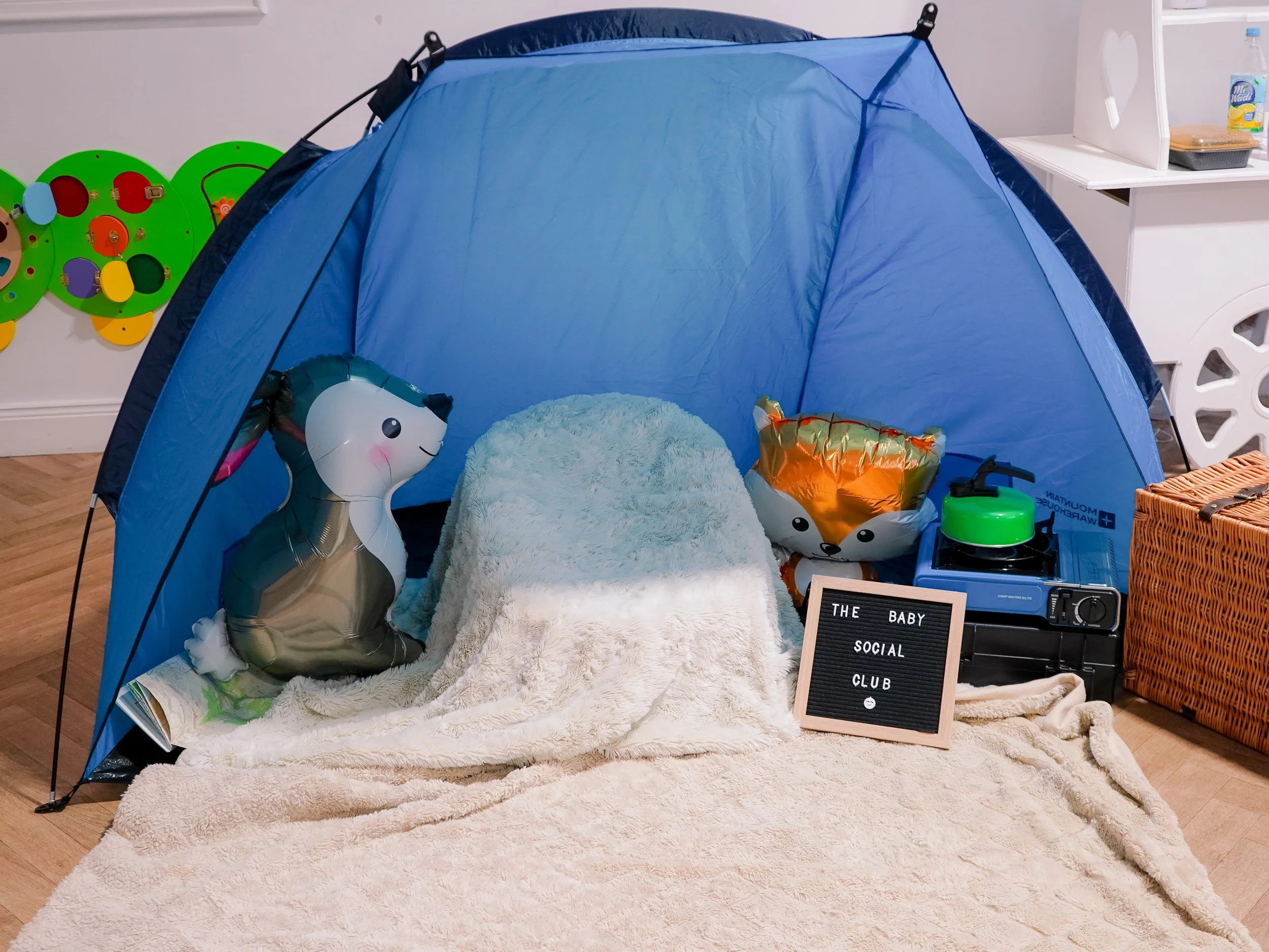 A small indoor camping setup with a blue tent on a cream-colored rug, decorated with animal-shaped balloons, including a unicorn and a fox. There is a letter board with the words 'THE BABY SOCIAL CLUB', a wicker basket, a play kitchen, and playful wall decorations in the background.