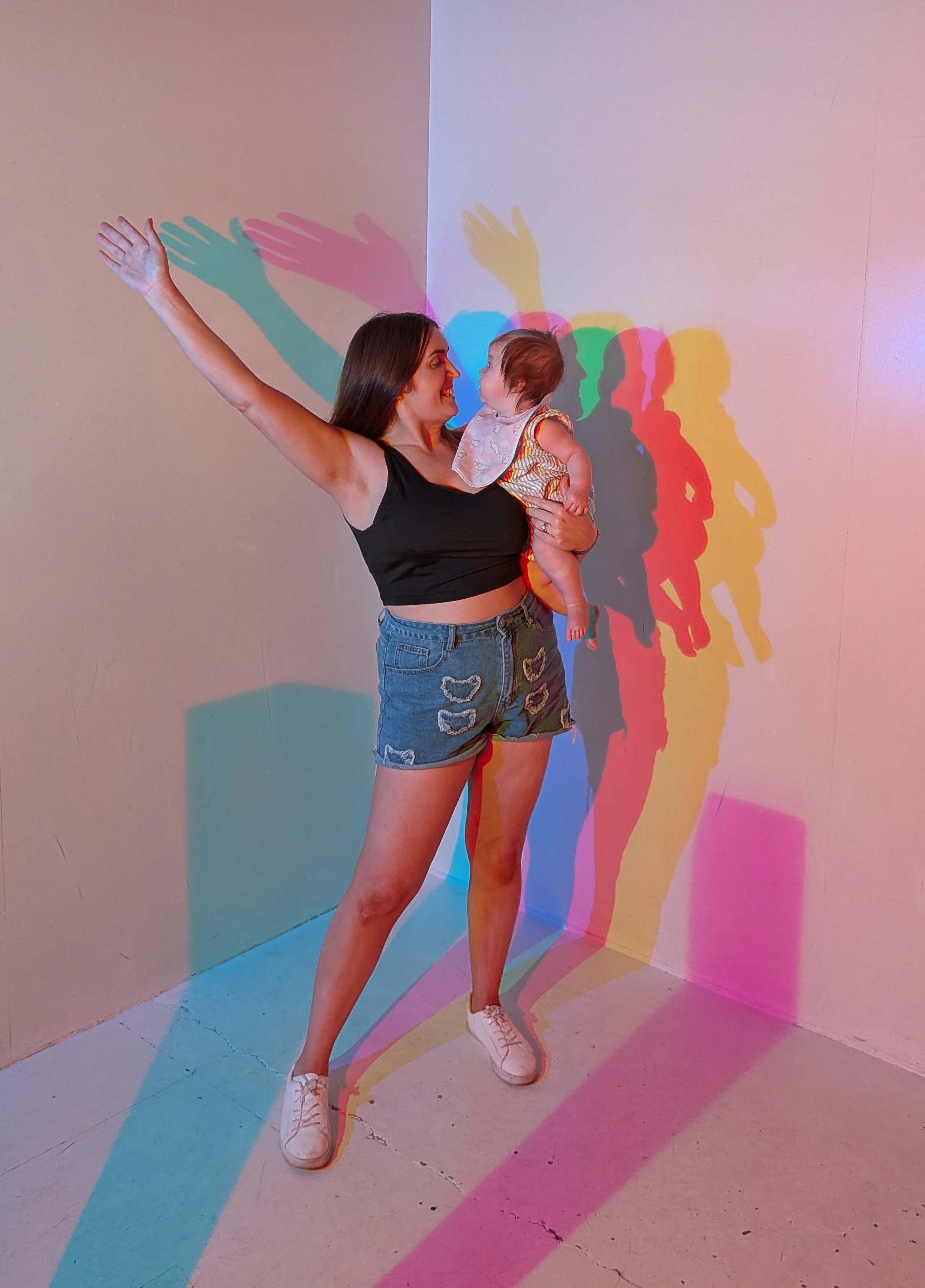 A woman with long dark hair wearing a black crop top, denim shorts with patches, and white sneakers holding a small child with short hair and a patterned dress, standing in a corner where colorful shadow projections of her raised arm and other figures are cast on the wall.