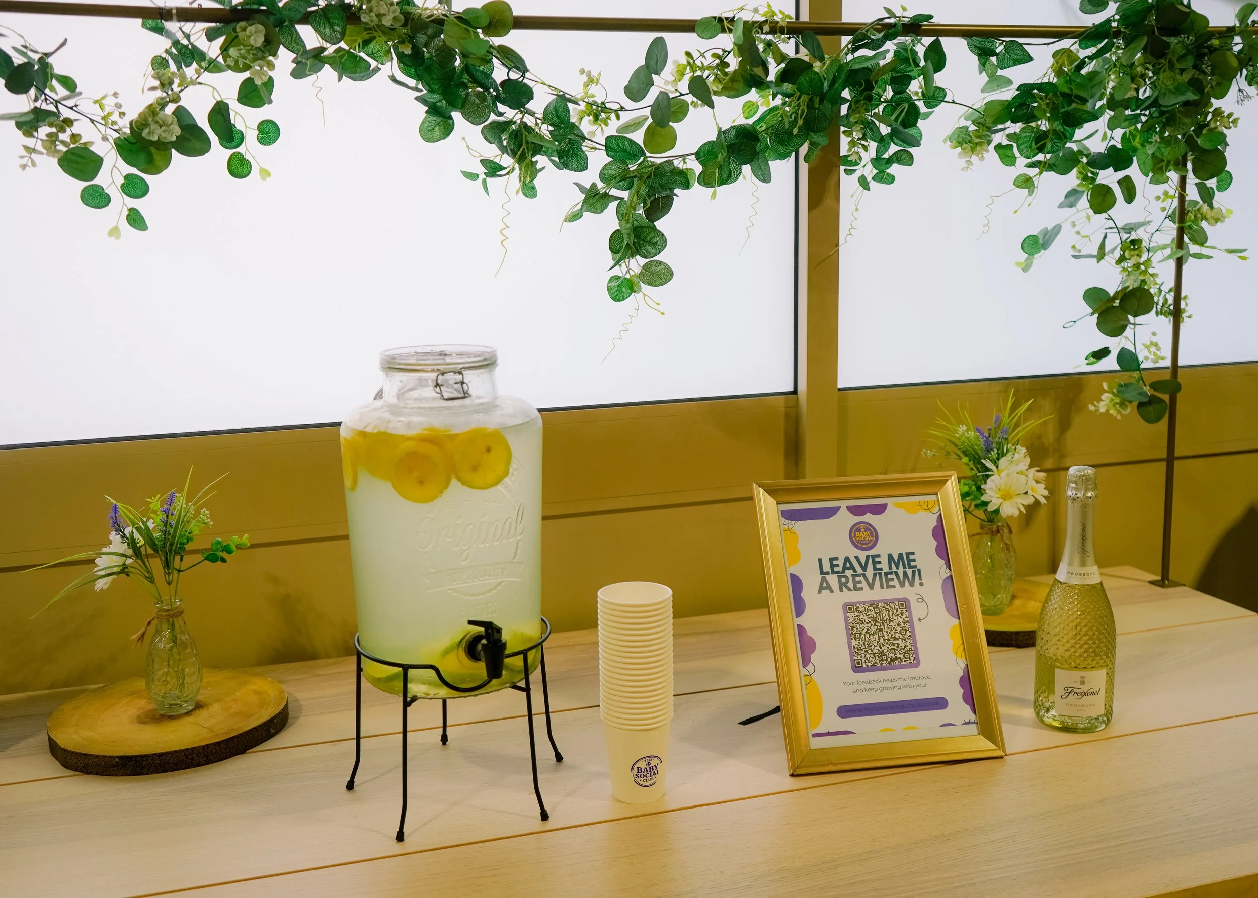 A display table with a large glass jar filled with lemonade and lemon slices, paper cups, a framed sign asking for reviews, and small floral arrangements, set in front of a backlit wall with greenery hanging.
