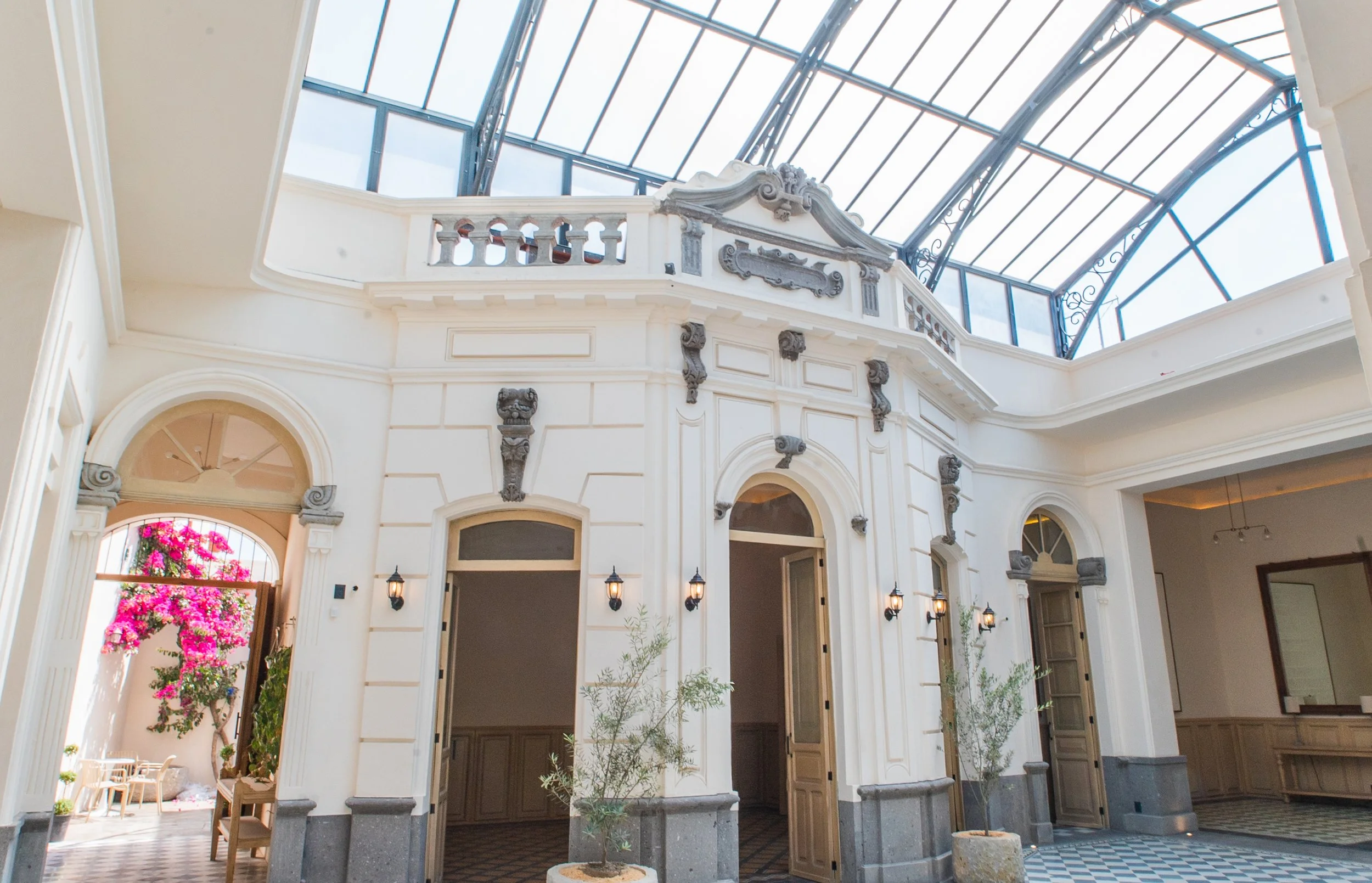 Interior de un edificio con techo de vidrio y estructura de metal, paredes blancas con detalles arquitectónicos decorativos, puertas abiertas, plantas y lámparas de pared.