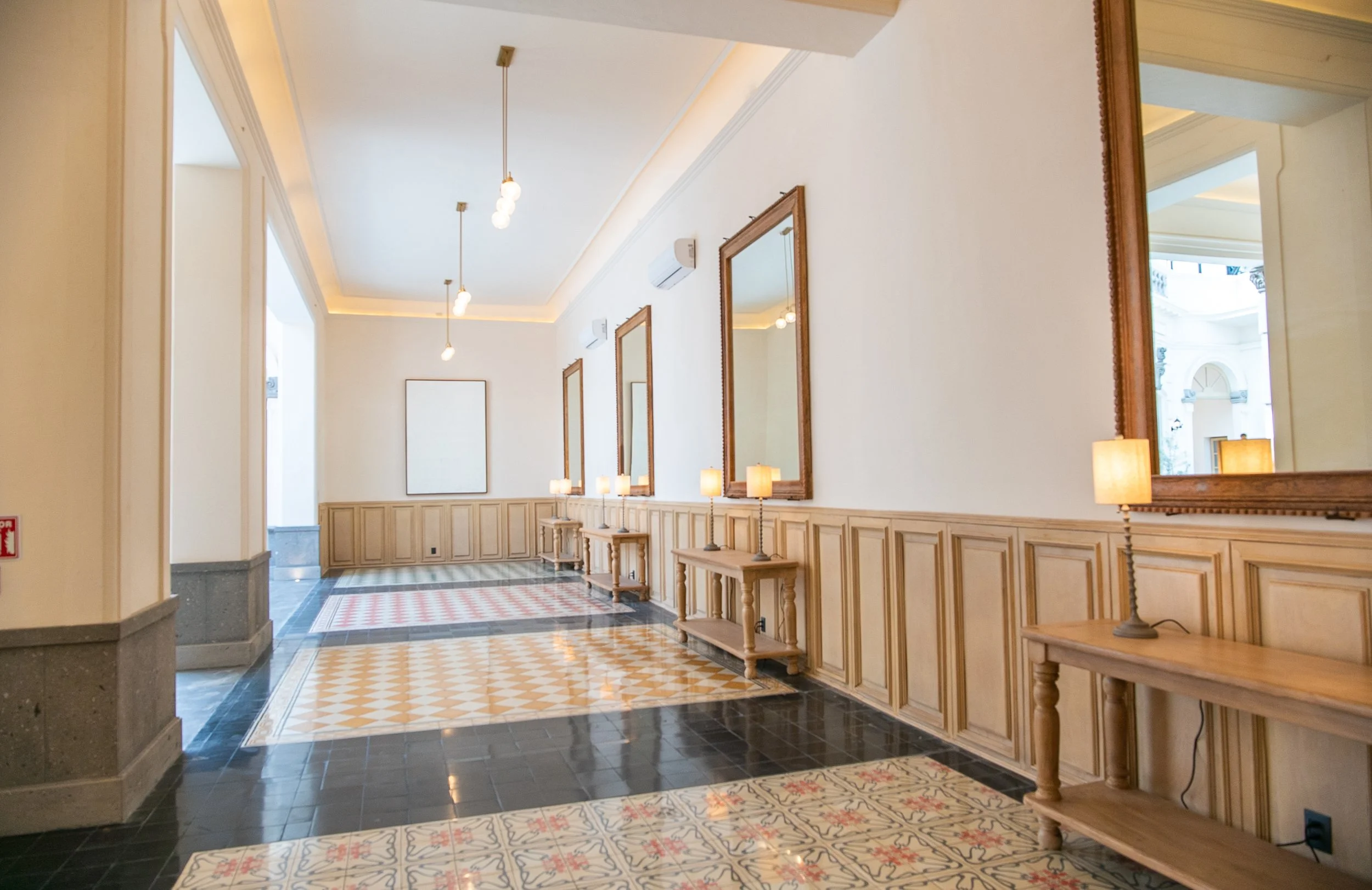 Vestíbulo con paredes blancas, espejos grandes con marcos de madera, lámparas de mesa sobre mesas de madera y un tapete con patrones coloridos en el piso.