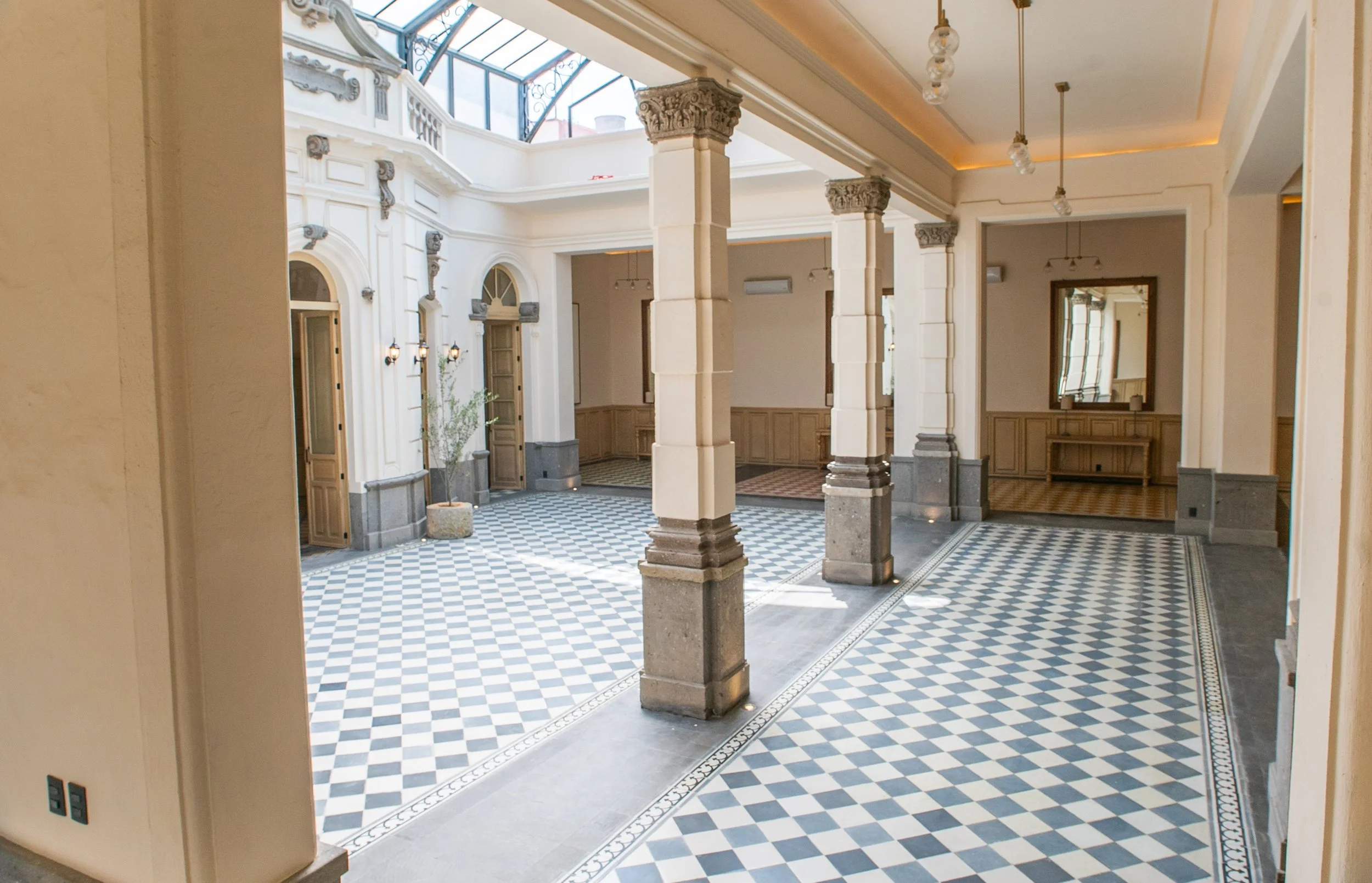 Interior de un edificio con columnas decorativas, paredes claras, techo con iluminación empotrada y un suelo de baldosas en patrón de cuadros diagonales en tonos azul y beige.