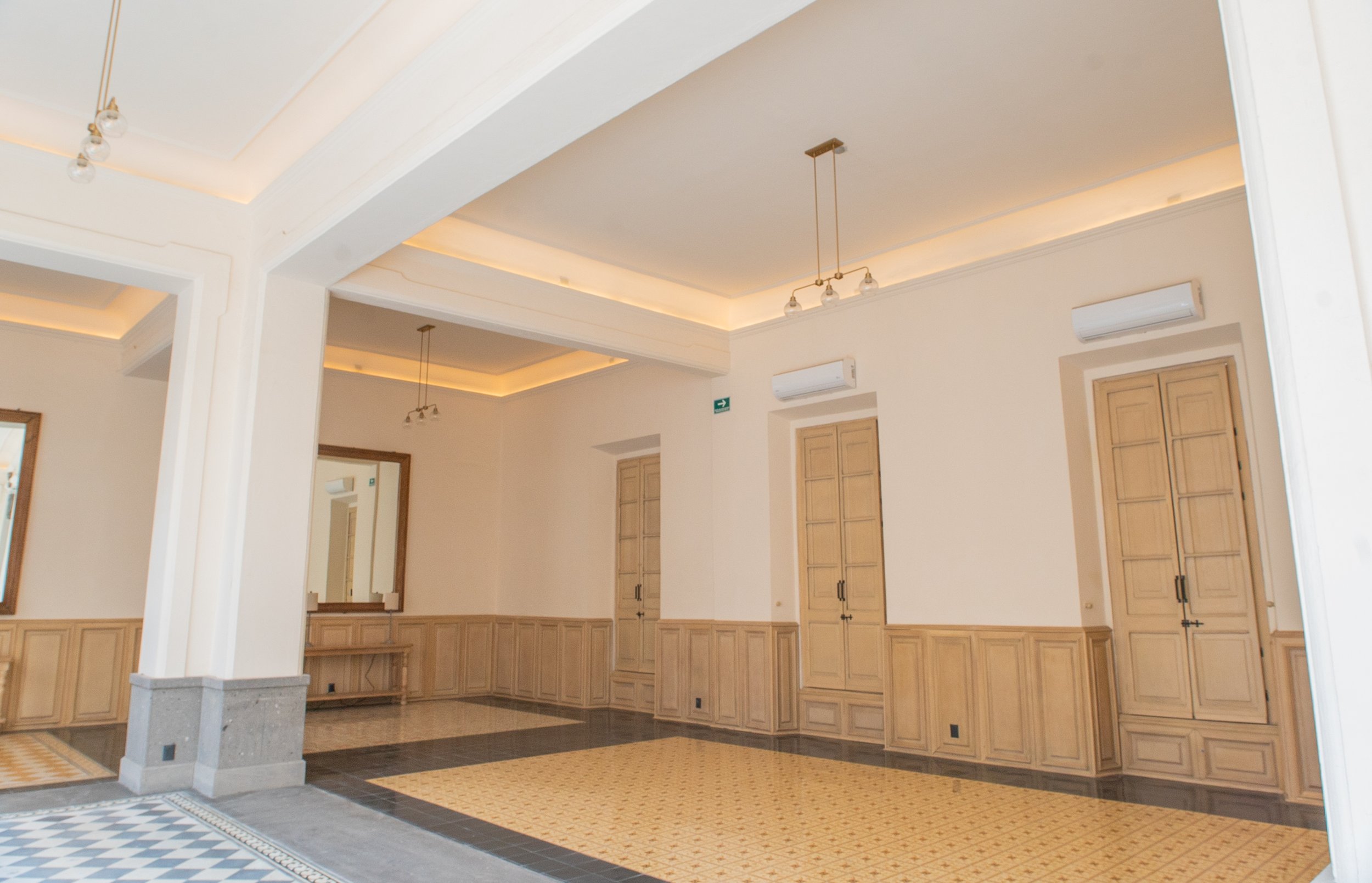 Sala amplia con paredes beige, puertas de madera, espejos en las paredes, aire acondicionado y cortinas blancas.
