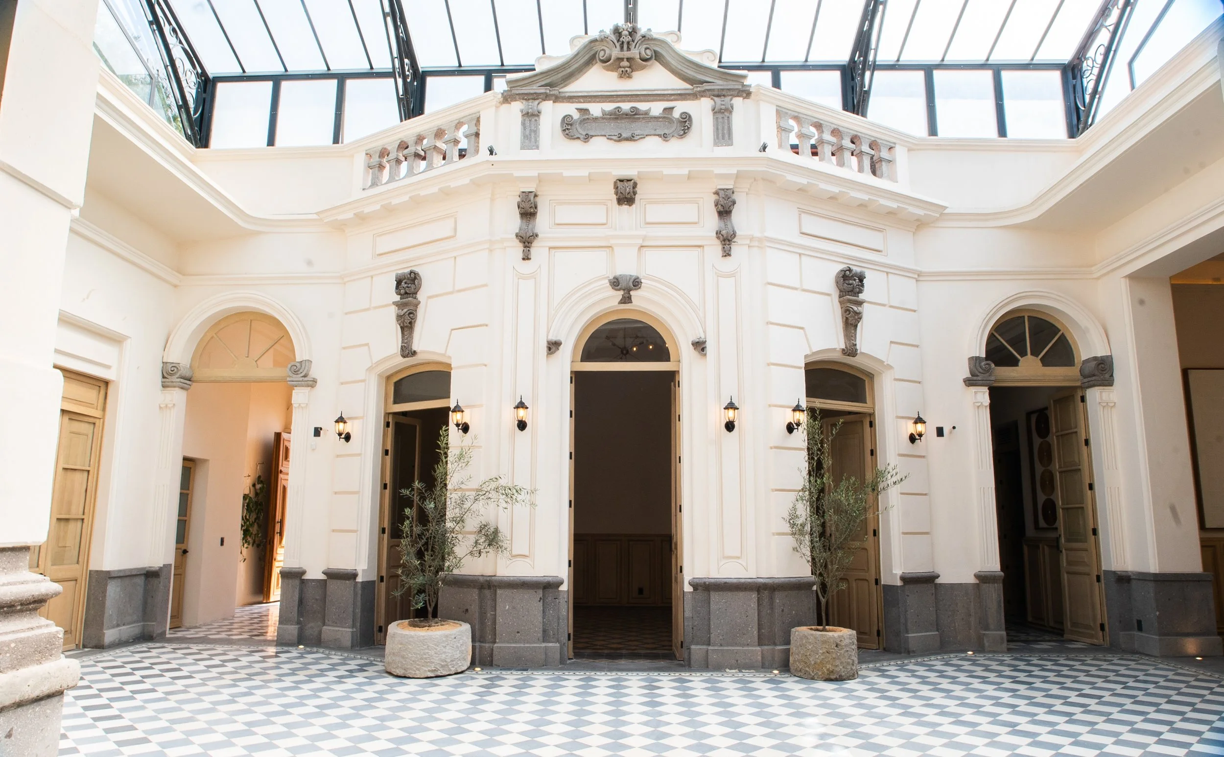 Interior de un edificio con arquitectura clásica, paredes blancas, puertas de madera, plantas en macetas, suelo de baldosas en patrón de ajedrez y un techo de cristal que deja entrar luz natural.