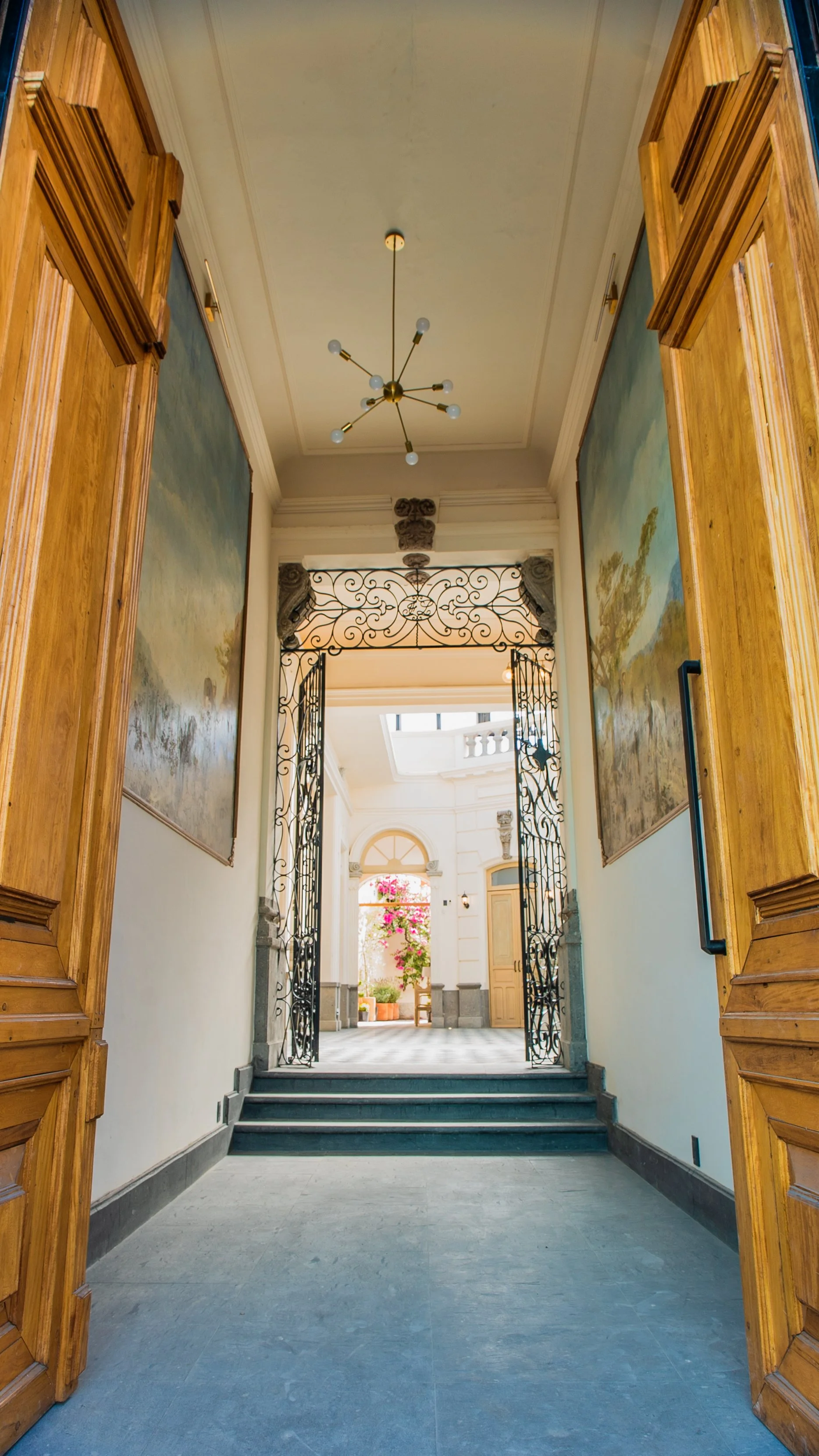Entrada elegante de un edificio con puertas de madera abiertas, paredes decoradas con murales, escaleras y una reja de metal, con un área interior luminosa y plantas en el fondo.