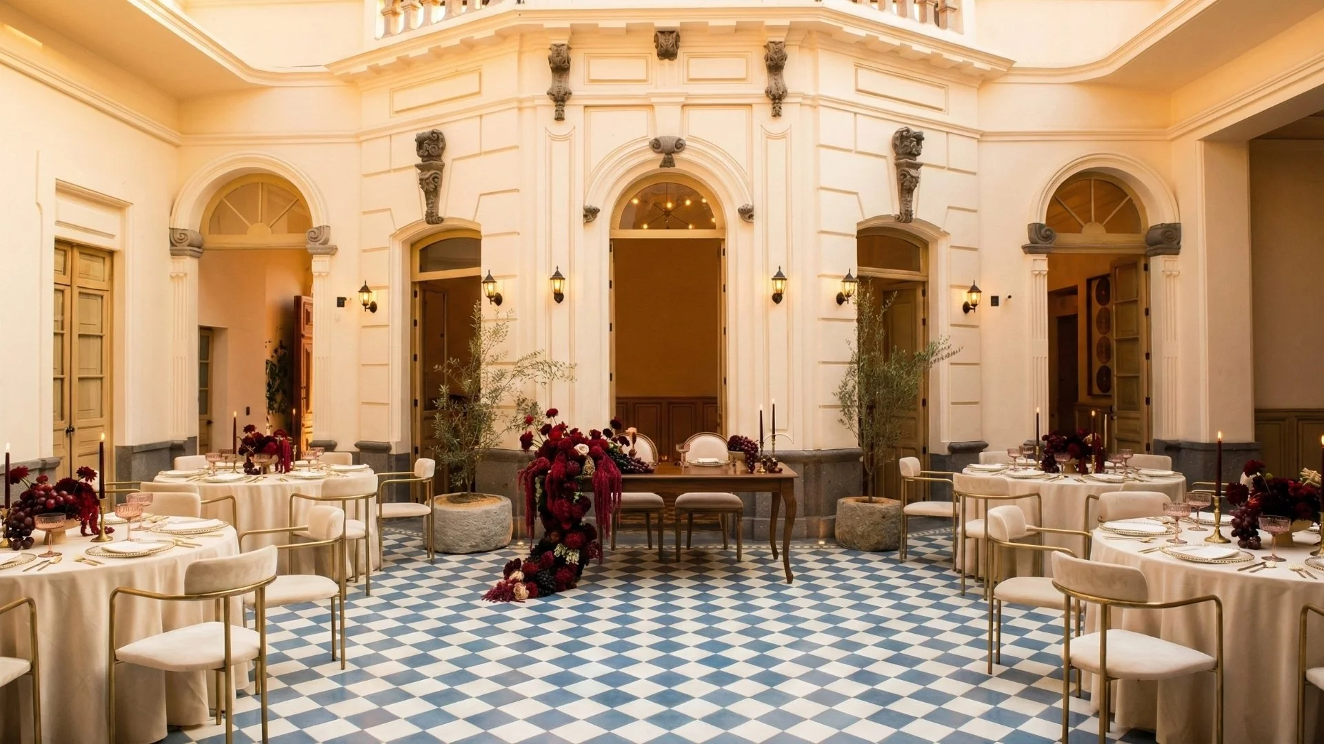 Salón elegante con mesas decoradas para una celebración, con manteles blancos, centros de flores rojas y candelabros, y paredes antiguas con molduras y puertas ornamentadas.