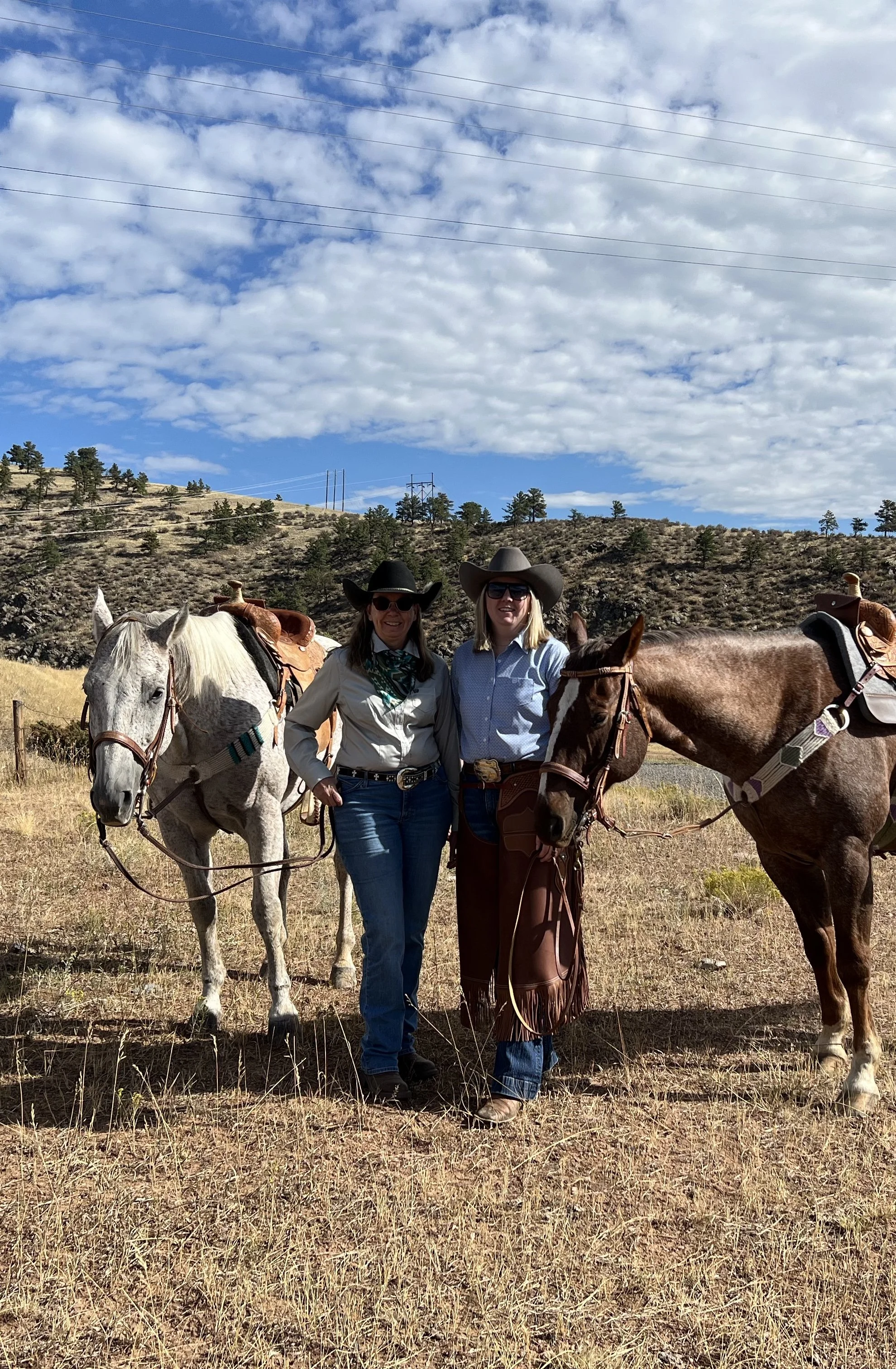 Two women standing outdoors in a dry, grassy field, each holding a horse, with hills and scattered trees in the background, wearing cowboy hats and western attire under a partly cloudy sky.