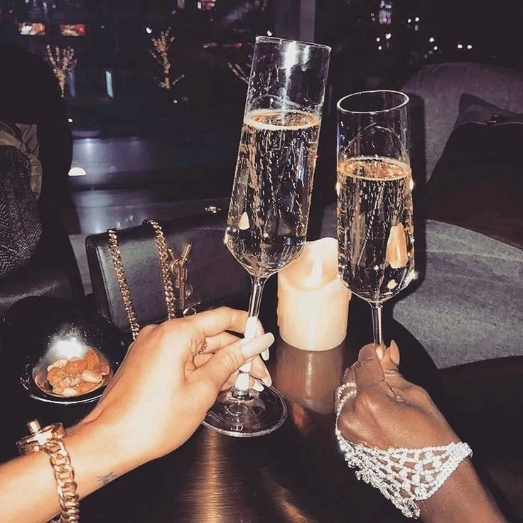 Two women holding glasses of champagne at a social gathering, with a candle and personal items on the table.