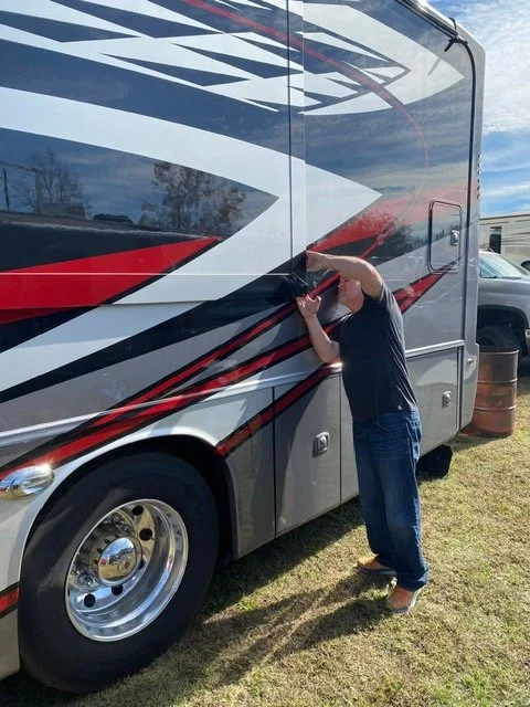 A person cleaning or working on the side of a large RV or bus with a gray, black, red, and white design, outdoors on grass near a building and a trash barrel.