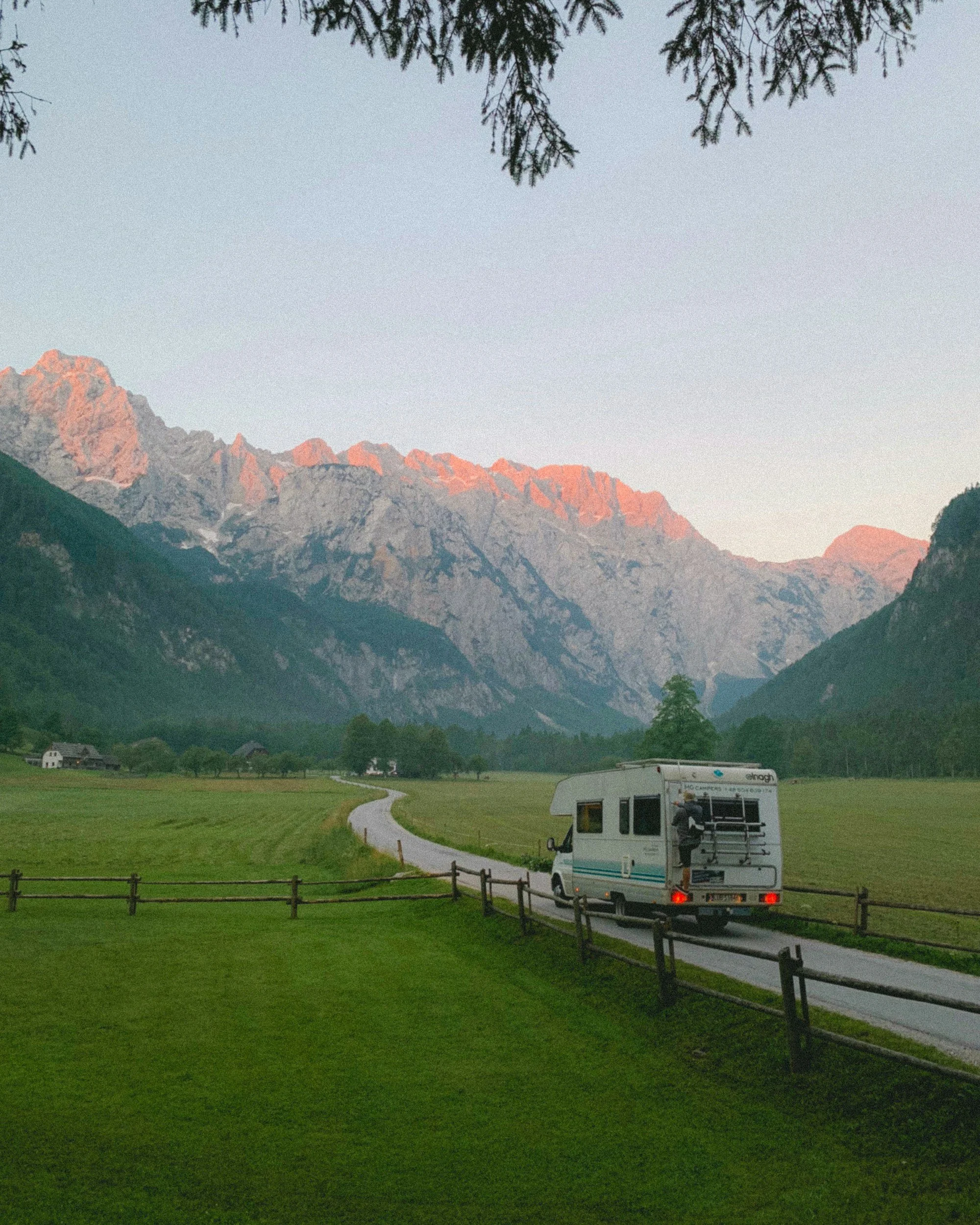 A scenic mountain landscape at dusk shows rocky mountain peaks with pink hues, surrounded by forested slopes. In the foreground, there is a green grassy field with a winding path, a small white building, and a camper van parked near a wooden fence. Overhead, tree branches frame the top of the image.