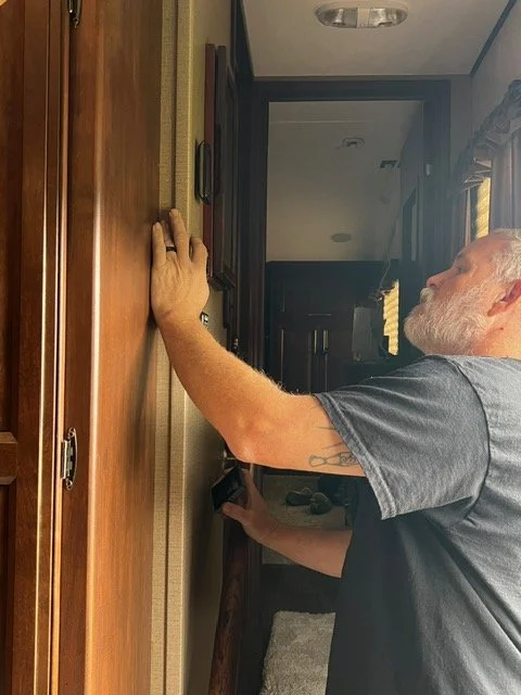 An older man with a white beard and gray hair is reaching up with his right hand towards a wooden cabinet or wall, using his left hand to hold what appears to be a tool or device. He is wearing a gray t-shirt and is in a room with a door, a window with curtains, and a carpeted floor.