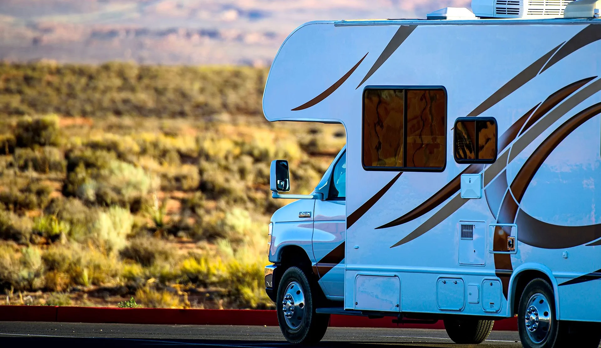 A white recreational vehicle with brown and black stripes parked on the side of a road amid a desert landscape with shrubs and a distant canyon.