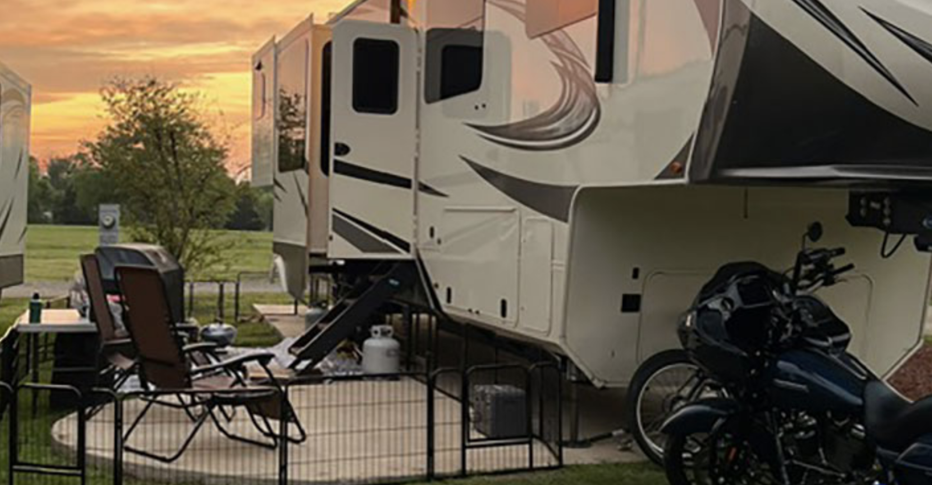 A white and black travel trailer parked on a lawn during sunset, with chairs, tables, a motorcycle, and a propane tank nearby, and a tree in the background.