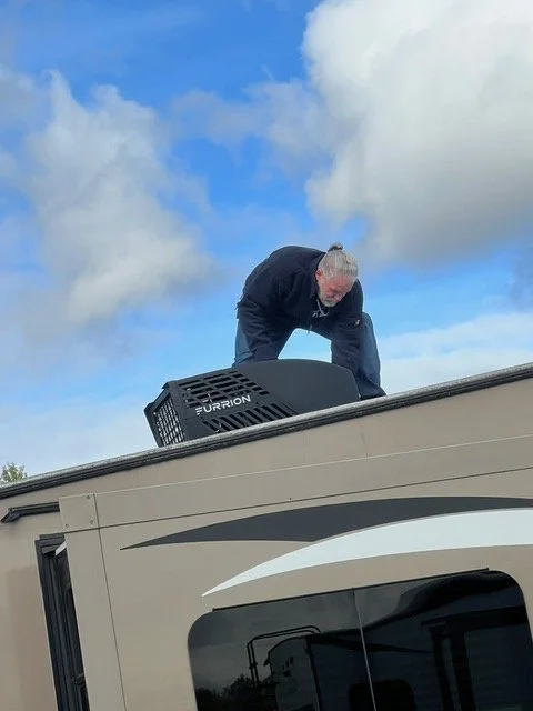 An elderly man with gray hair crouched on the roof of a vehicle near a pet carrier labeled 'Furrion' against a partly cloudy sky.
