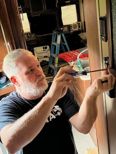 A man with a beard and gray hair fixing or installing something on a wall, using a screwdriver, inside a room with various tools and equipment.