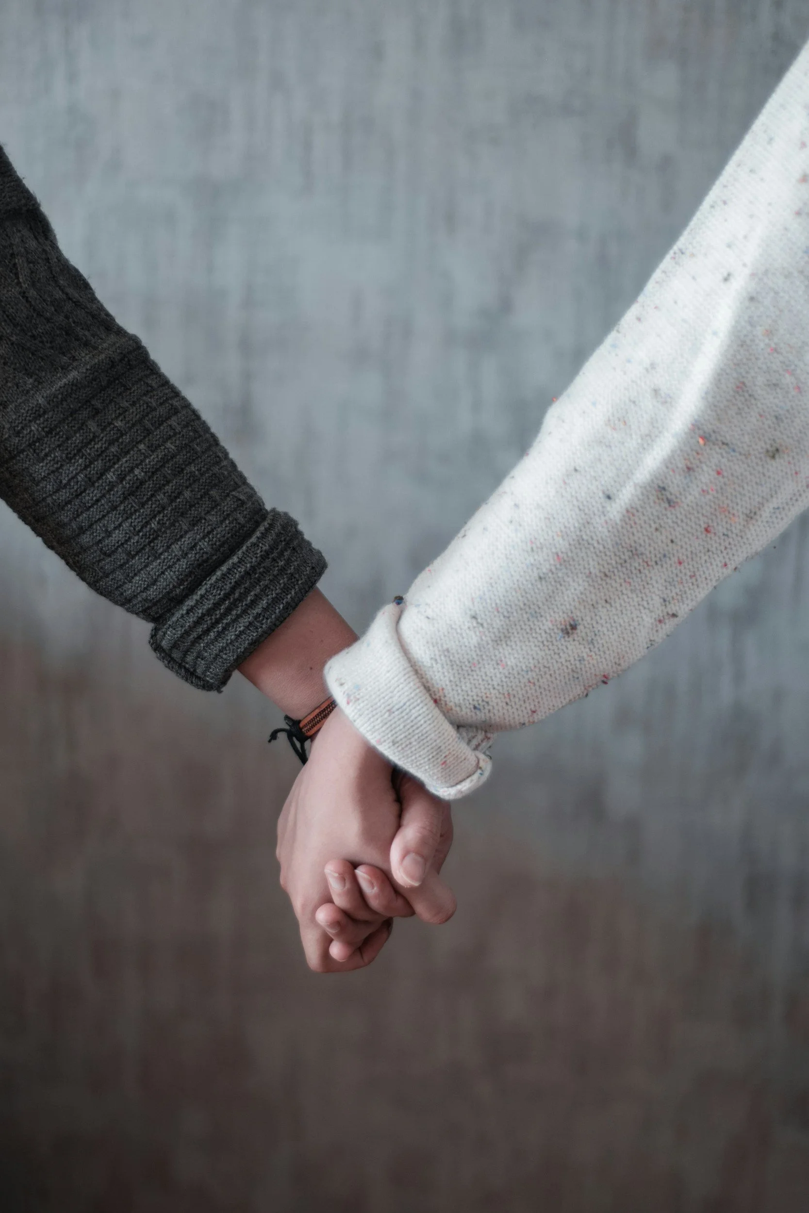 Two people hold hands. Both people are wearing long sleeves. There is a neutral colored background.