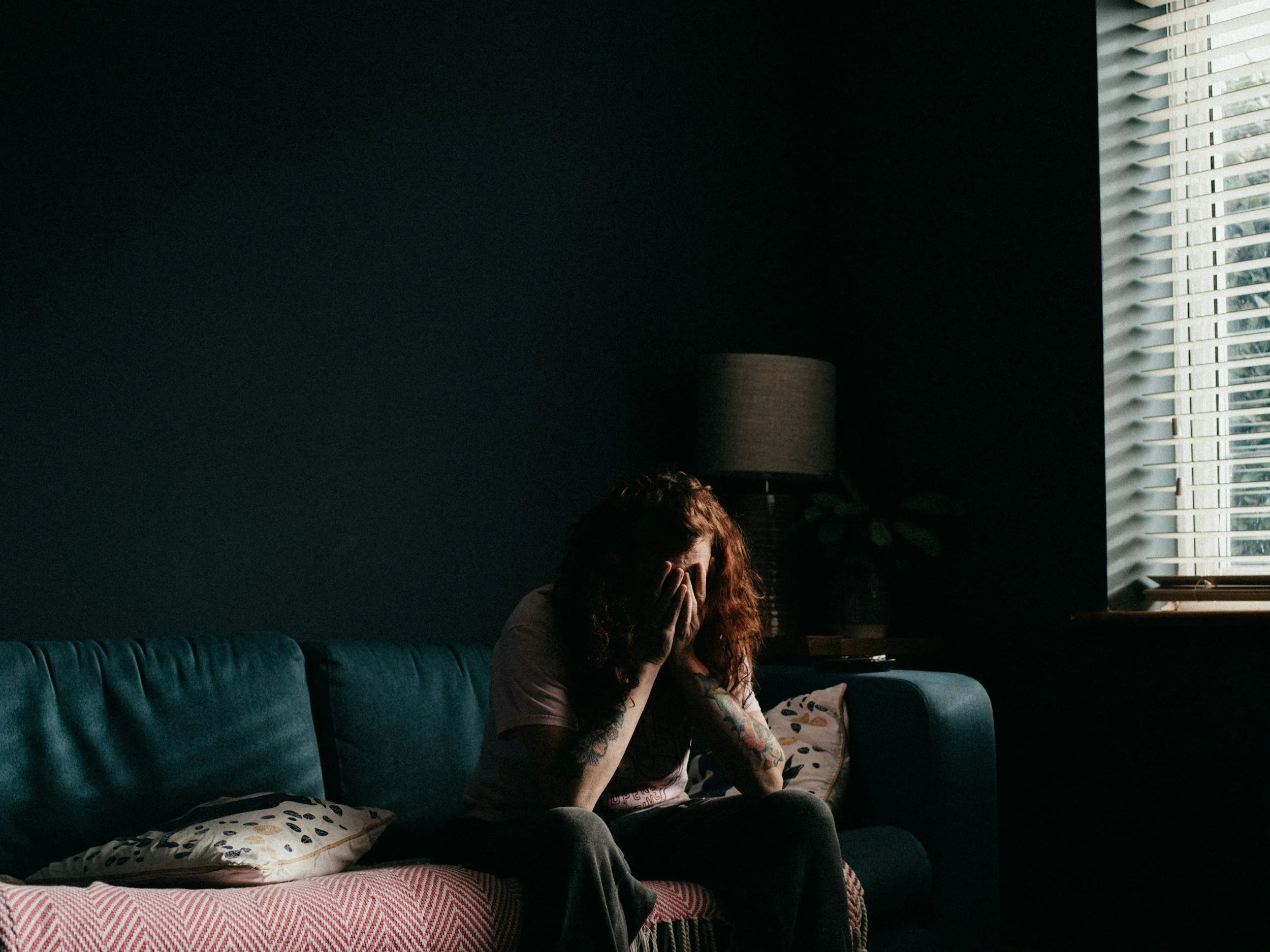 A woman sits on her couch with her face in her hands, appearing overwhelmed or sad. She is wearing comfortable athletic clothing. The lamp behind her is off and there is a small amount of light coming from a window.