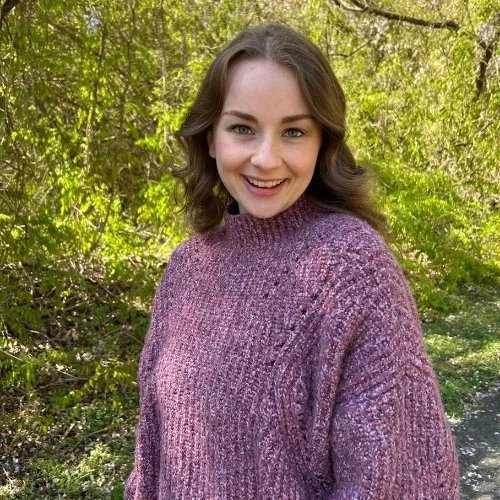 Headshot of Amelia. She is wearing a pink sweater and is standing in a natural area with greenery behind her.