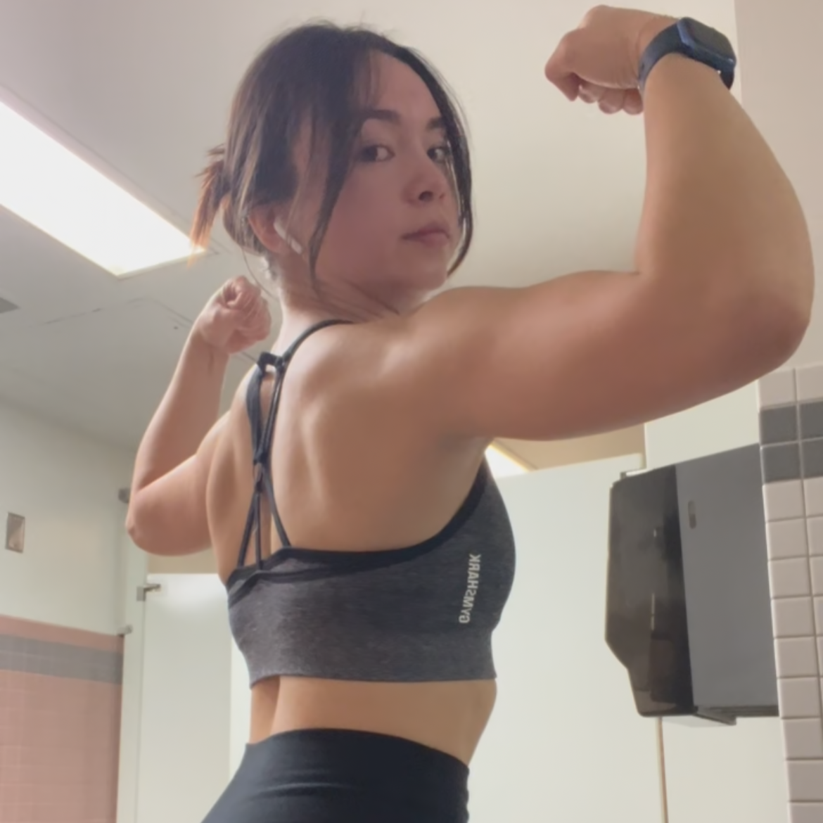 Woman flexing her arm muscles in a gym bathroom mirror.