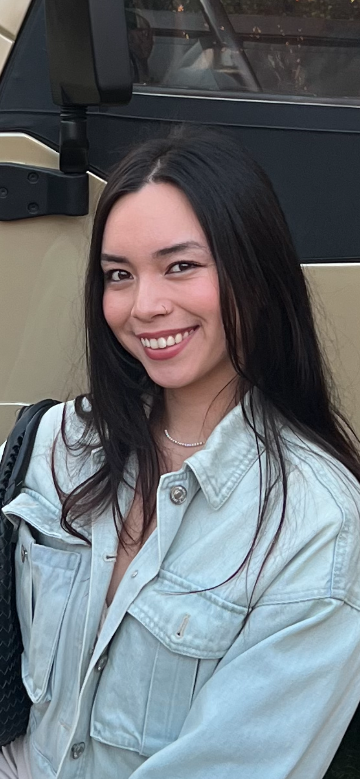 A woman with black hair, smiling, wearing a light denim jacket and a pearl necklace, standing outdoors near a vehicle.