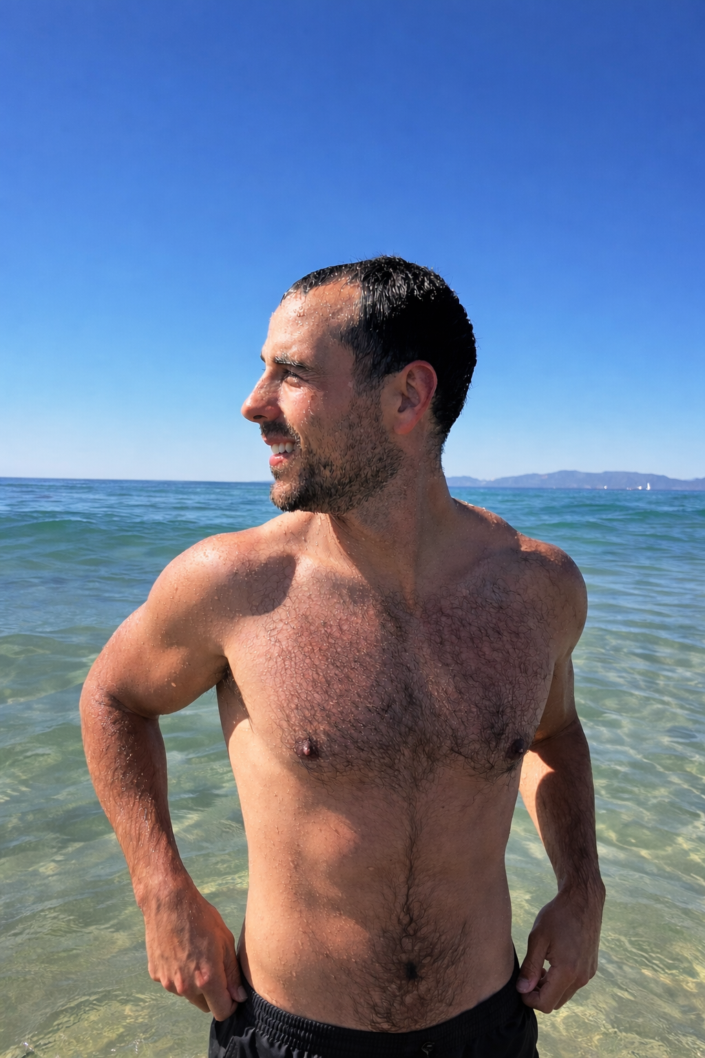 A man with wet hair and a beard standing shirtless at the beach, looking to the side, with a clear blue sky and ocean in the background.