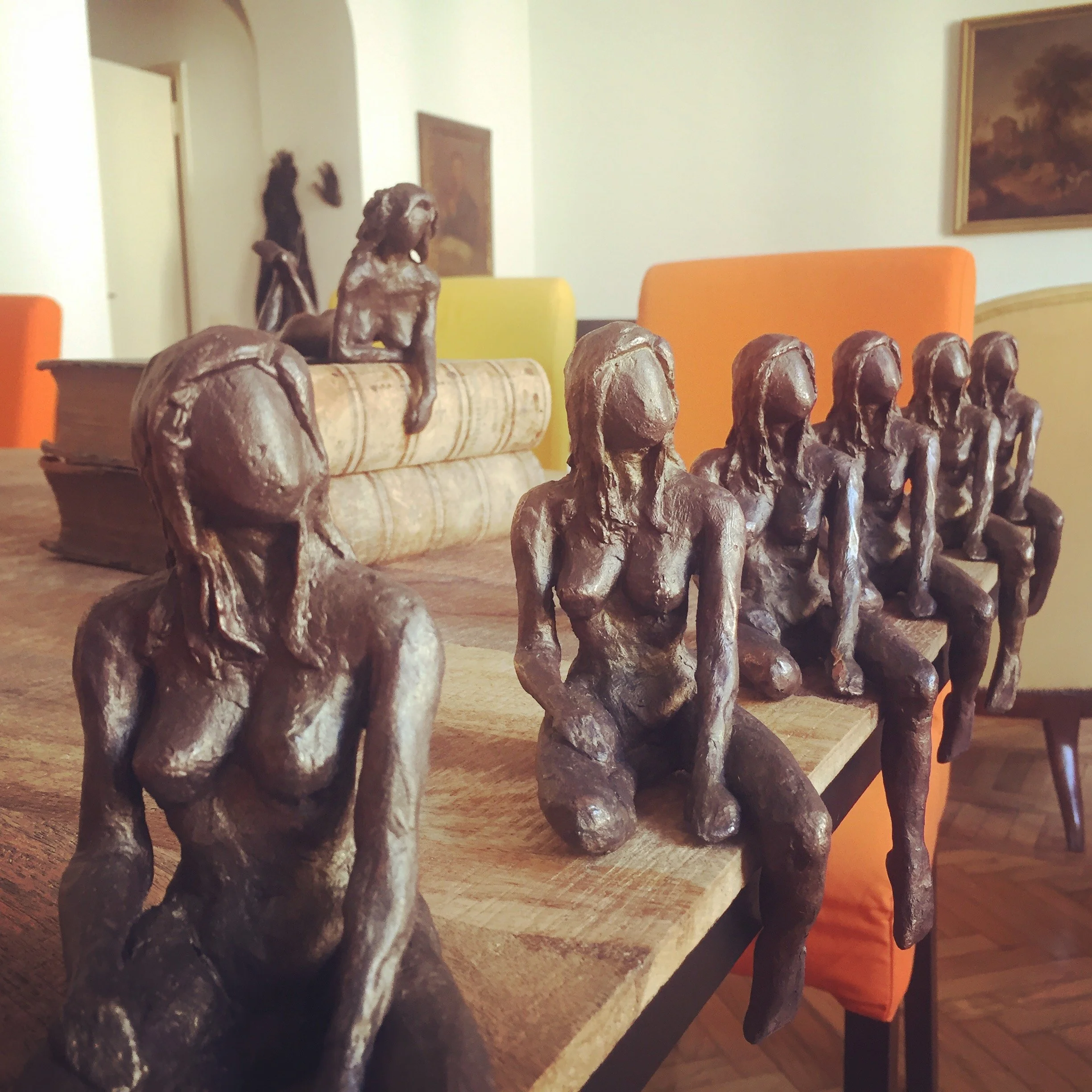 Statue di donne senza gave, disposte su un tavolo in legno, con uno sfondo di pareti bianche, una libreria, un quadro e sedie di diversi colori.