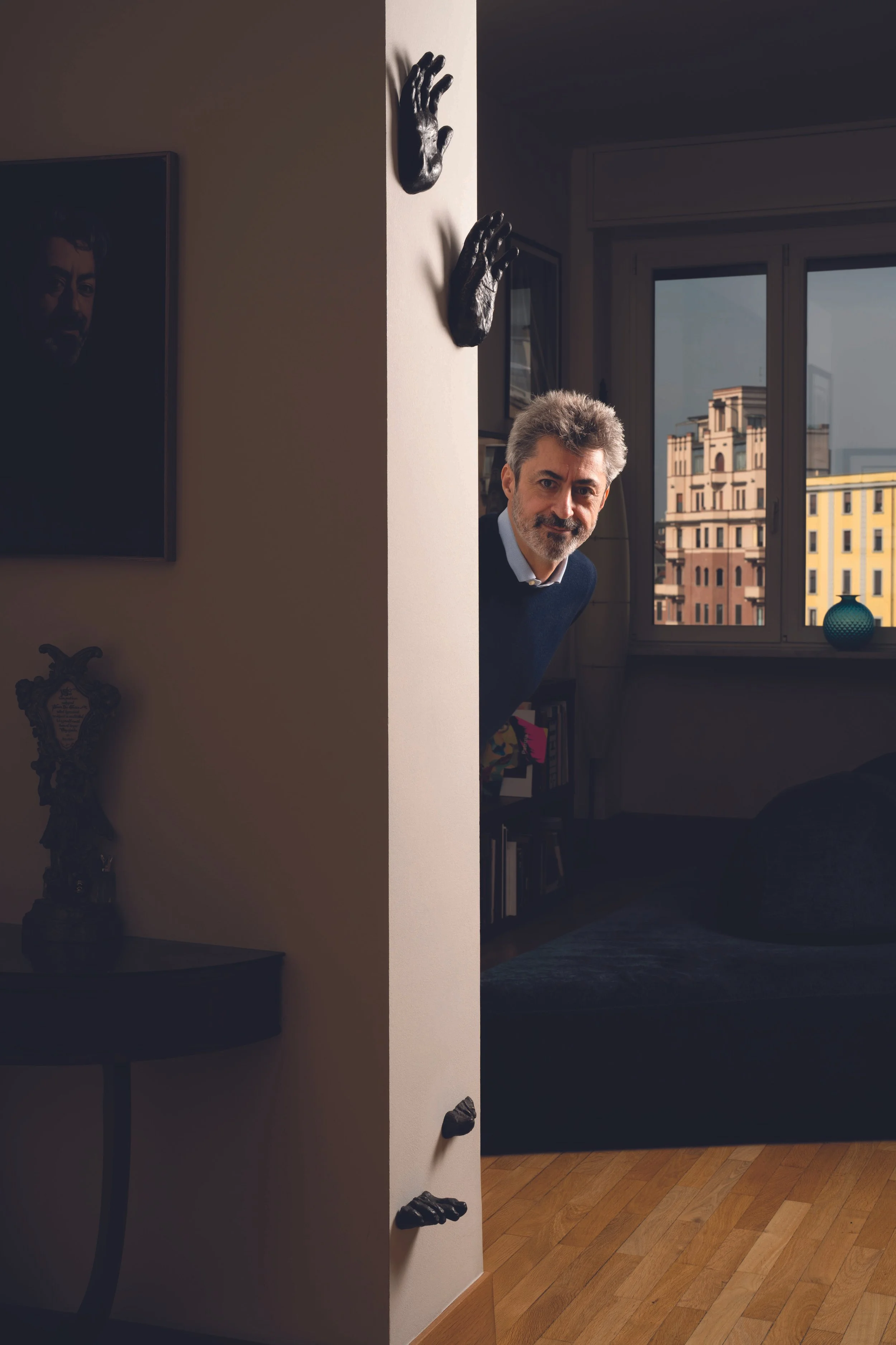 Uomo con capelli grigi e barba che sporge da dietro una parete in una stanza ben arredata, con pareti decorate e grandi finestre, in un ambiente luminoso.