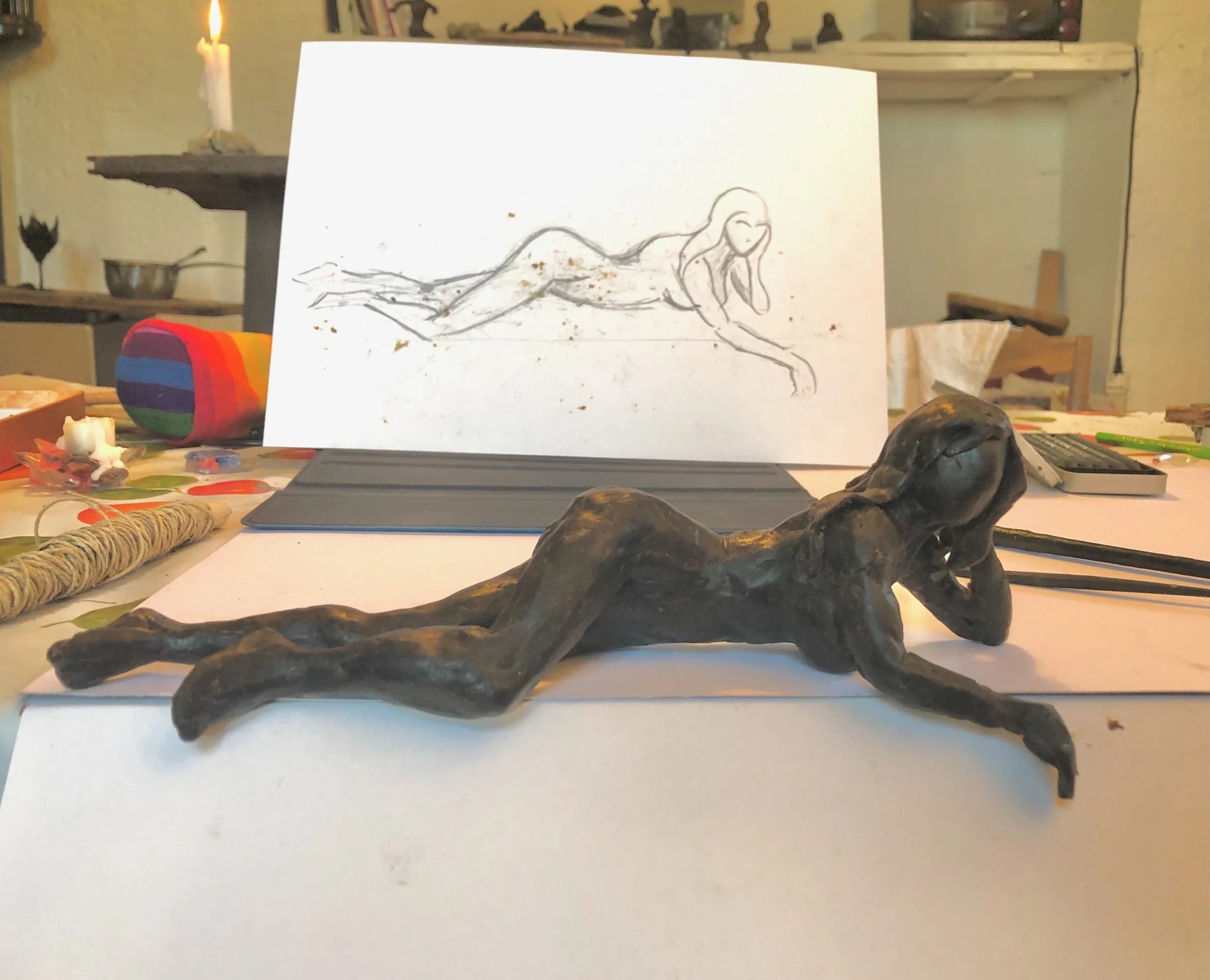 Statua in bronzo o ceramic a forma di donna sdraiata con la testa appoggiata alla mano, collocata su un tavolo. Sullo sfondo, un quadro con disegno di una stessa forma umana. Vari strumenti artistici e materiali sono sparsi sul tavolo.