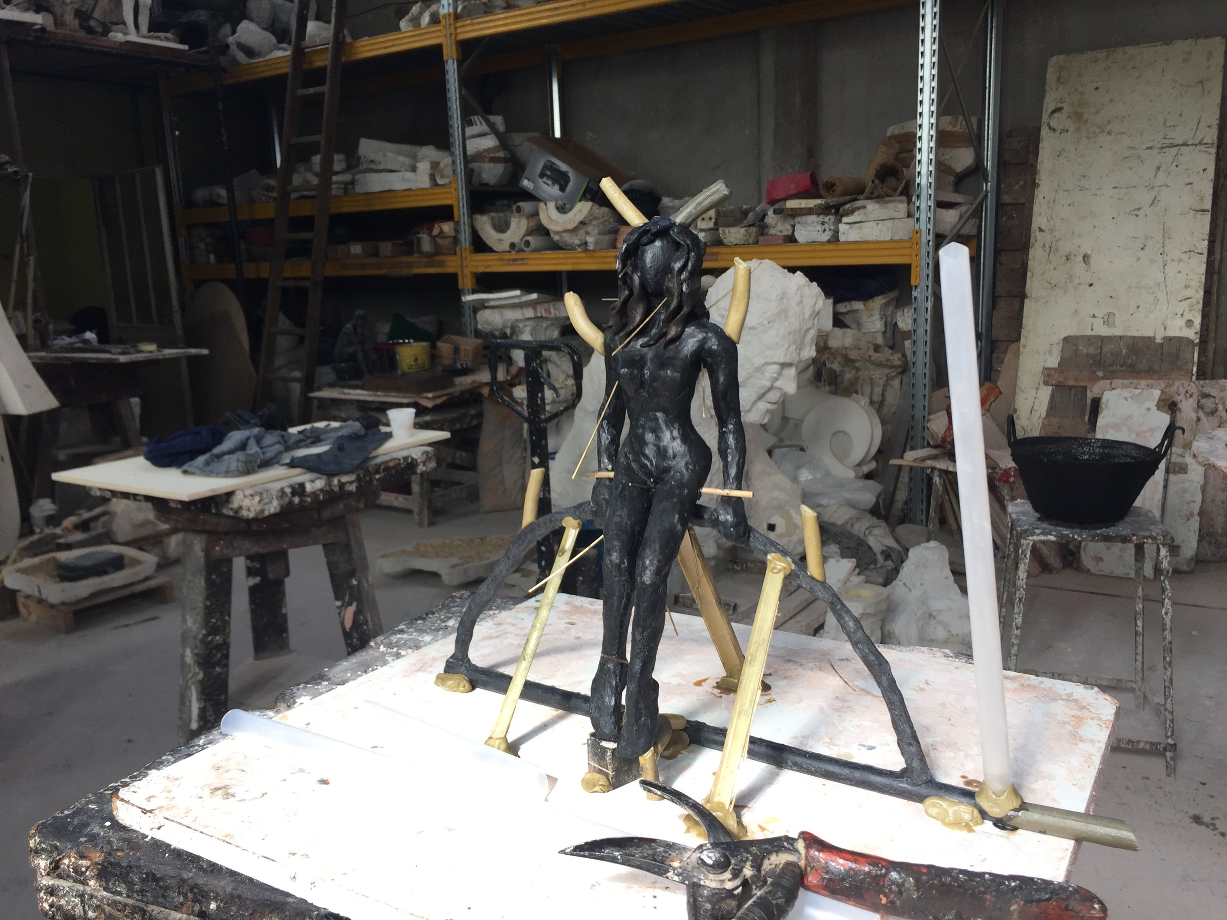 Statua in corso di lavorazione di una figura femminile, probabilmente di un angelo o di una dea, con elementi di supporto e strumenti artistici nella sua fase di creazione in uno studio di scultura.