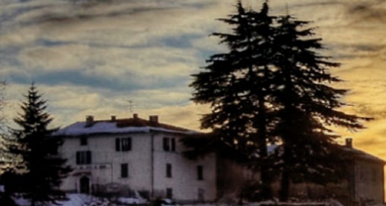Casa bianca con tetto rosso e due grandi pini in primo piano, cielo nuvoloso al tramonto.