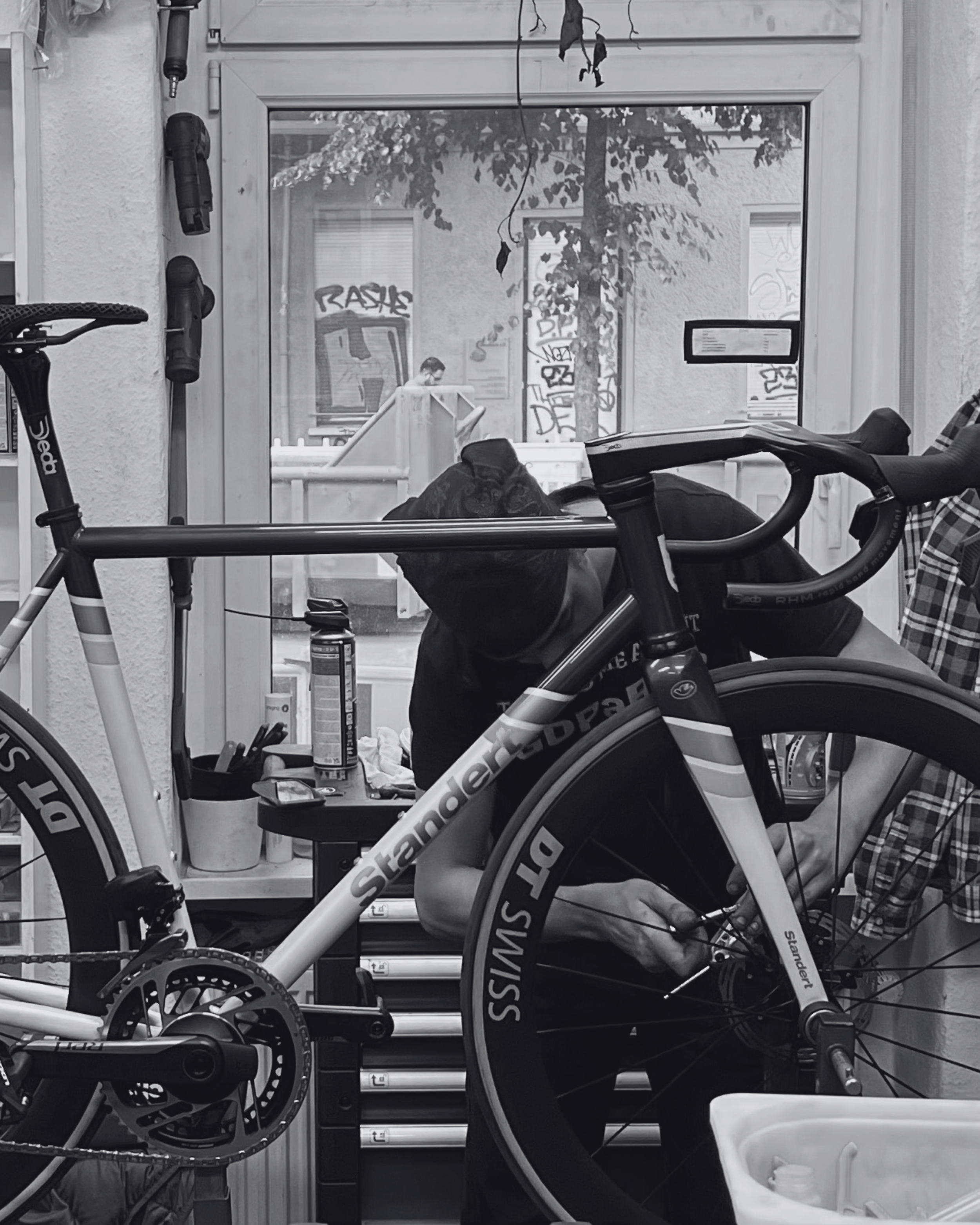 A person working on a bicycle inside a workshop, with tools and supplies around them, seen through a window.