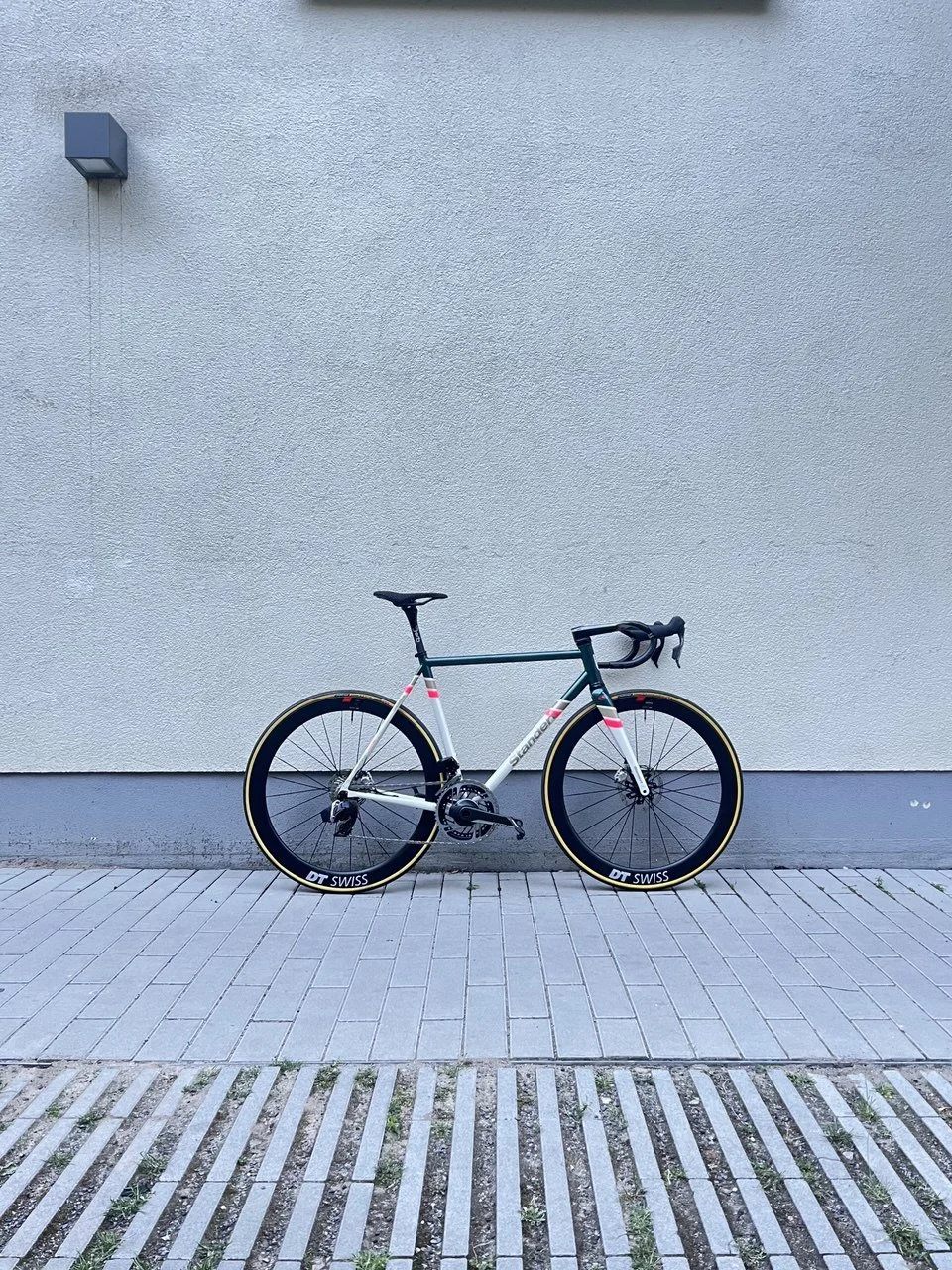 A modern road bicycle with thin tires and drop handlebars, leaning against a plain gray wall with a small wall-mounted light fixture nearby.