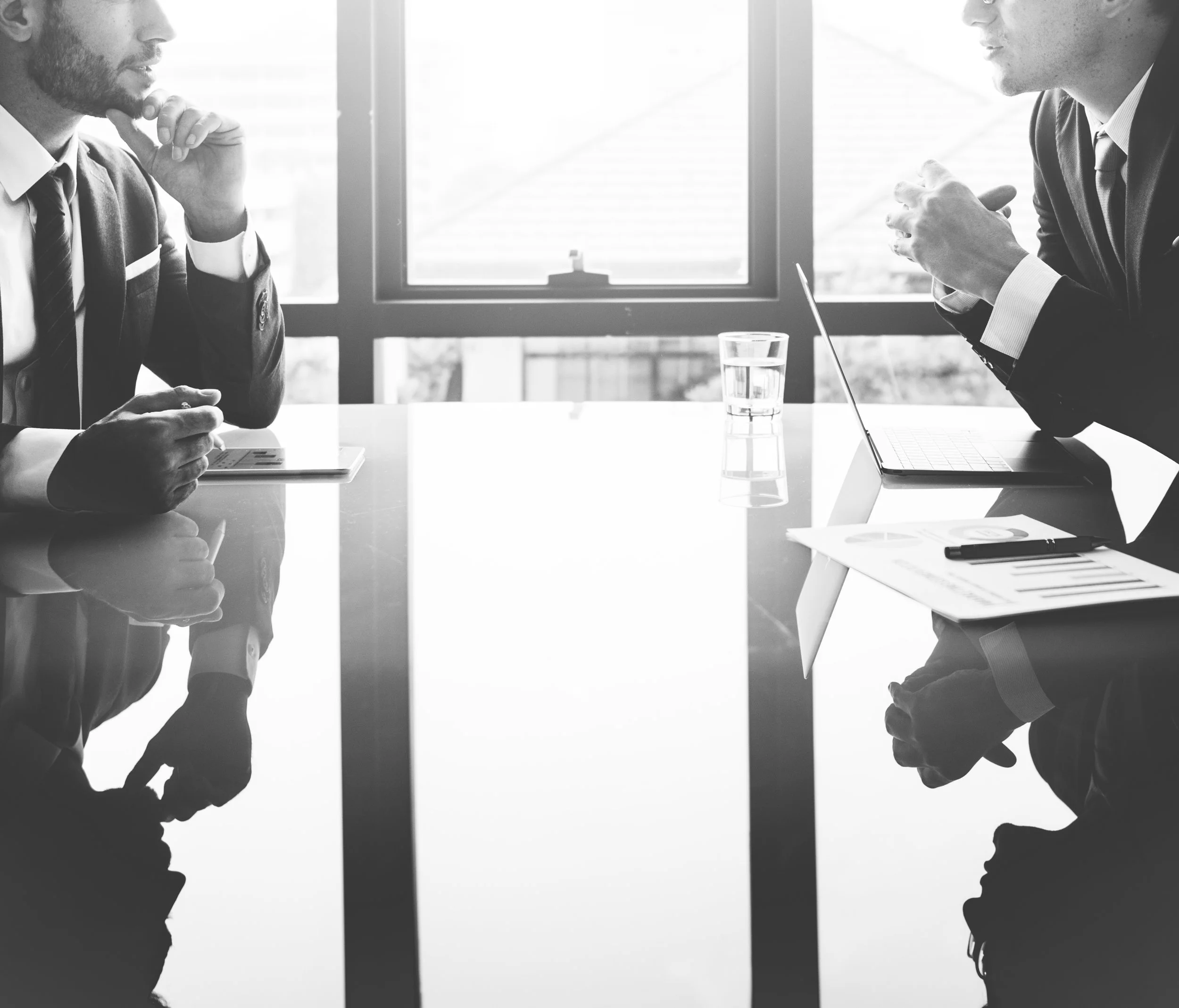 Two men in business suits having a discussion in a modern office with large windows, a glass of water, a laptop, and documents on the table.